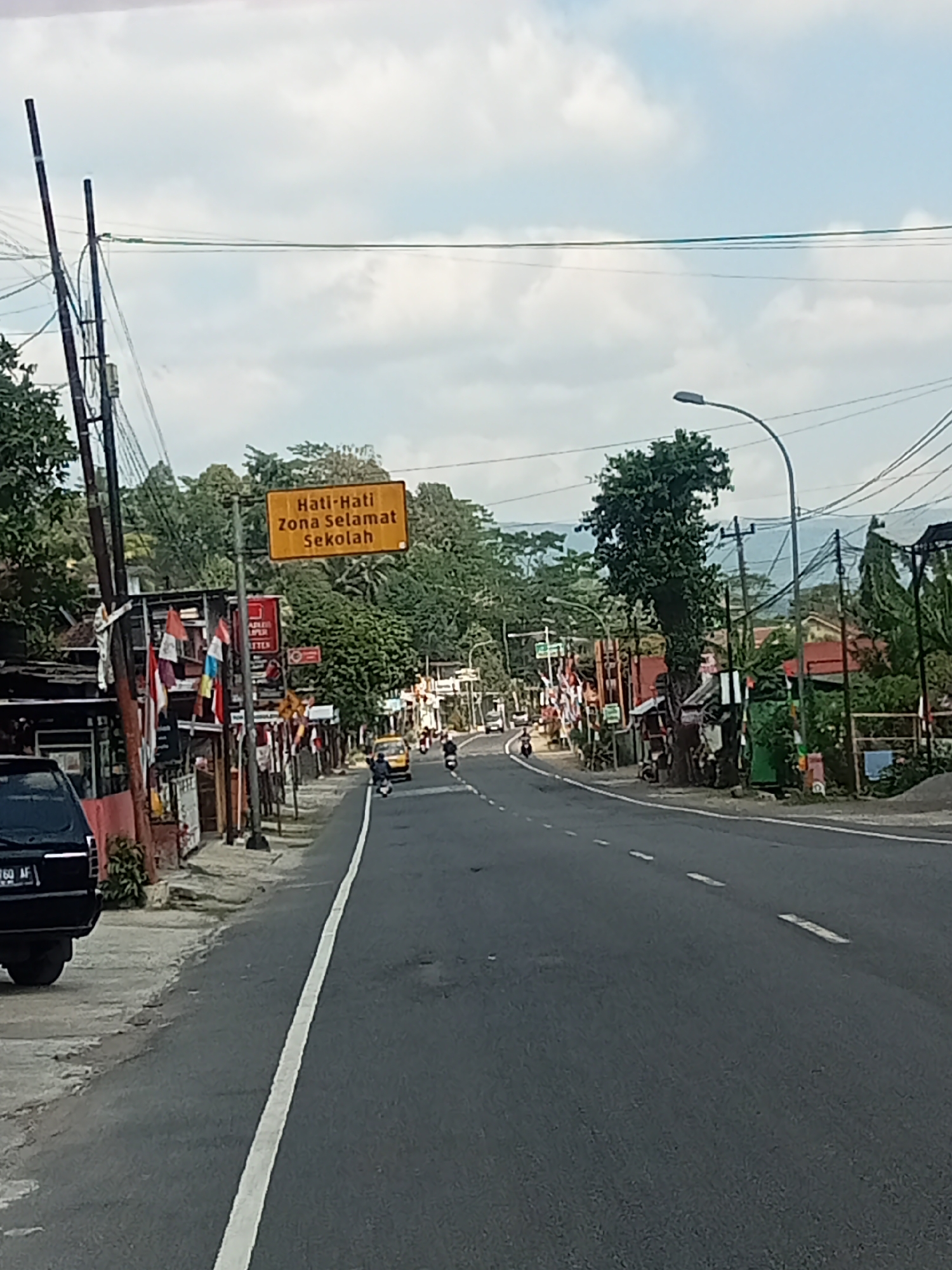 The image captures a street scene in Selokromo, Indonesia, likely during daylight hours under an overcast sky. The foreground shows a paved road with a white dividing line. To the left of the road, a black vehicle is parked. Further along the left side, there are buildings with awnings and numerous Indonesian flags displayed. A prominent yellow sign with black text reads "Hati-Hati Zona Selamat Sekolah," which translates to "Beware, Safe School Zone." Street poles with electrical wires are visible throughout the scene. In the middle ground, a yellow vehicle, possibly a bemo or a small bus, is driving on the road, along with several motorcycles. The background reveals a lush green, hilly landscape. The overall environment suggests a busy street in a town or village.