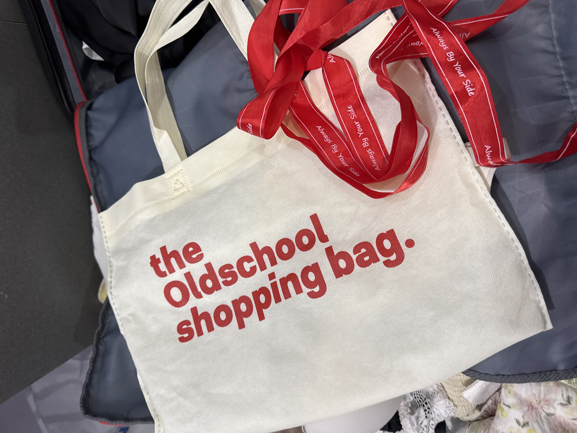 A white tote bag with the text "the Oldschool shopping bag." printed in red is partially visible, surrounded by items. A few red ribbons with "Always By Your Side" printed on them are on top of the bag. Other items, including what appear to be clothes and a zippered bag, are also visible, creating a cluttered scene. The location and city in Vietnam (Đà Nẵng) are not determinable from the image.