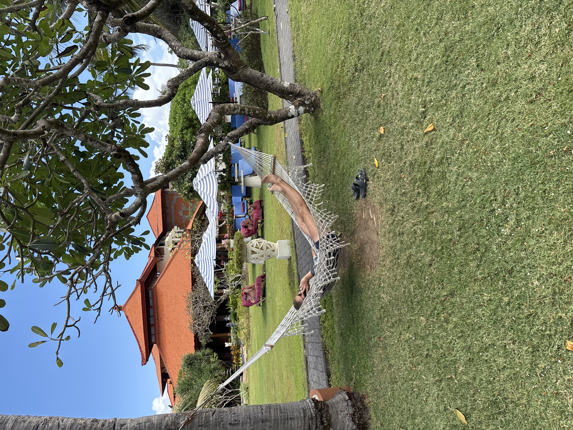 A person reclines in a hammock tied to a palm tree and another tree. The person is wearing sunglasses and dark swim trunks. The hammock is made of white netting. Black sandals are on the grass next to the hammock. The hammock is in a grassy area with a paved pathway behind it. In the background, there are several blue lounge chairs with striped umbrellas. Beyond the chairs, there is a large building with an orange tile roof and other structures with similar roofing. Trees and greenery are scattered throughout the scene. The sky is blue with some white clouds. This scene appears to be a resort or hotel in Kuta Selatan, Indonesia.