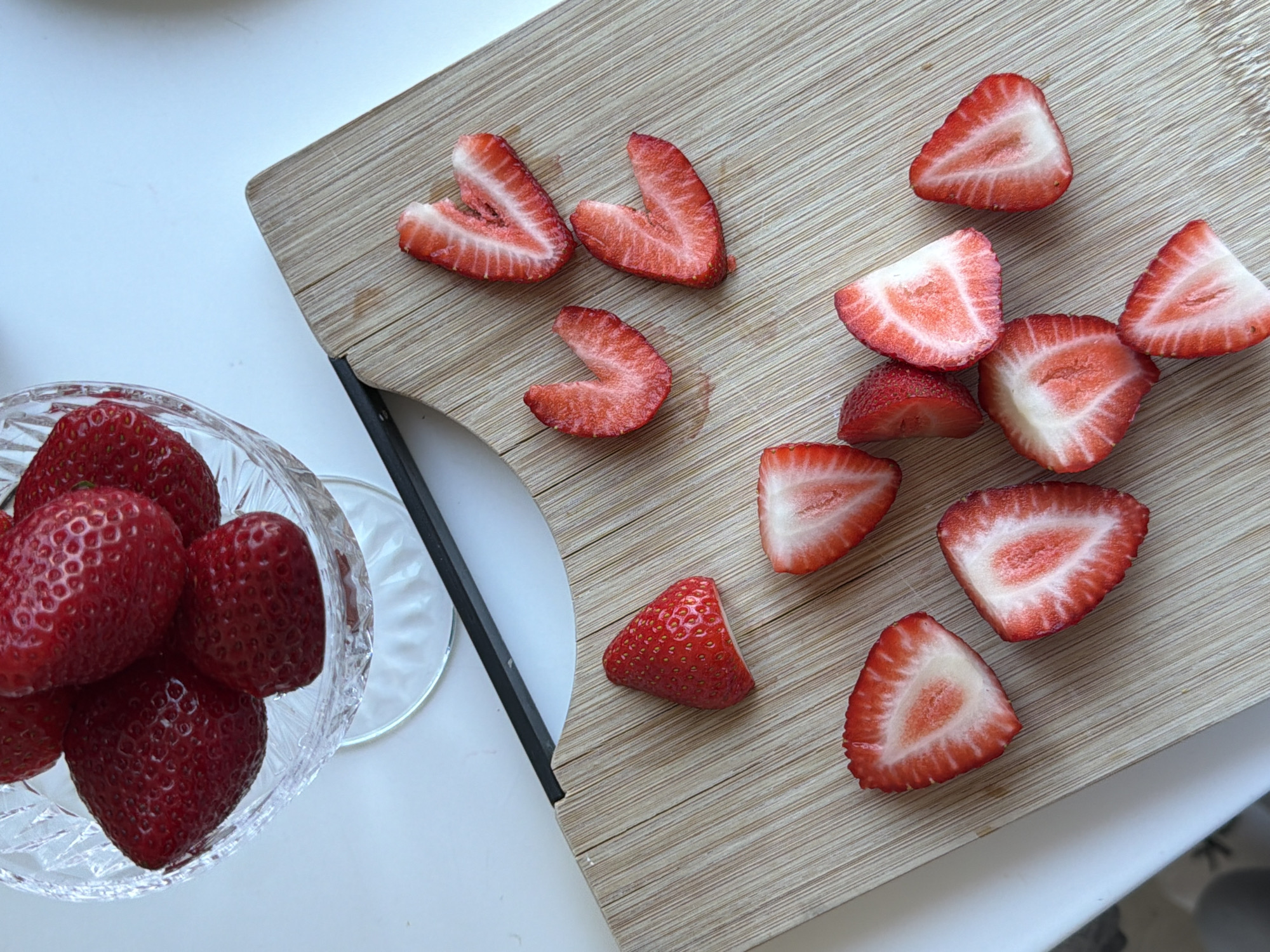 The image shows a top-down view of a clear crystal bowl filled with whole strawberries, placed on a white surface next to a light-colored wooden cutting board. The cutting board holds several sliced strawberries, mostly cut in half or into smaller pieces. A black object, possibly a knife or other tool, lies diagonally between the bowl and the cutting board. The environment is clean and well-lit, suggesting an indoor setting. There is no indication of the city of Dreiliņi, Latvia. No people, actions, or interactions are visible.