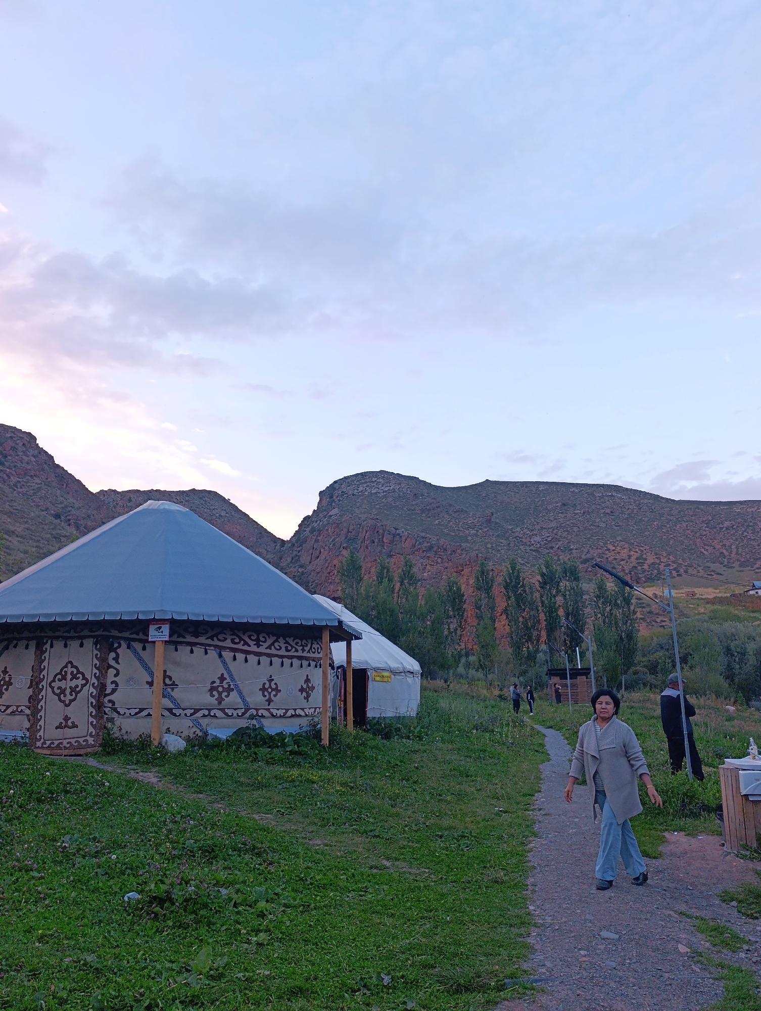 The image shows a scene in Kazan-Kuygan, Kyrgyzstan, with yurts set up in a grassy area at the base of a rocky mountain range. The sky is a pale blue with soft clouds, indicating either early morning or late evening.

In the foreground, a woman in a grey cardigan and blue pants is walking on a gravel path. Her expression is neutral, and she is looking towards the viewer. To her right, a man in a dark jacket and cap stands with his back to the camera.

Several yurts, traditional nomadic dwellings, are visible. The closest yurt is large with a conical roof and is decorated with intricate patterns. A sign with what appears to be a security camera icon is affixed to the yurt. Another white yurt is partially visible behind it.

The environment is a mix of natural and developed elements. Lush green grass covers the ground, dotted with small purple flowers. Trees line the middle ground, and the imposing, barren mountains form the backdrop. A basic wooden structure, possibly a restroom or information booth, is situated to the right of the path. Several other indistinct figures are visible further down the path, suggesting this is a tourist or communal area.