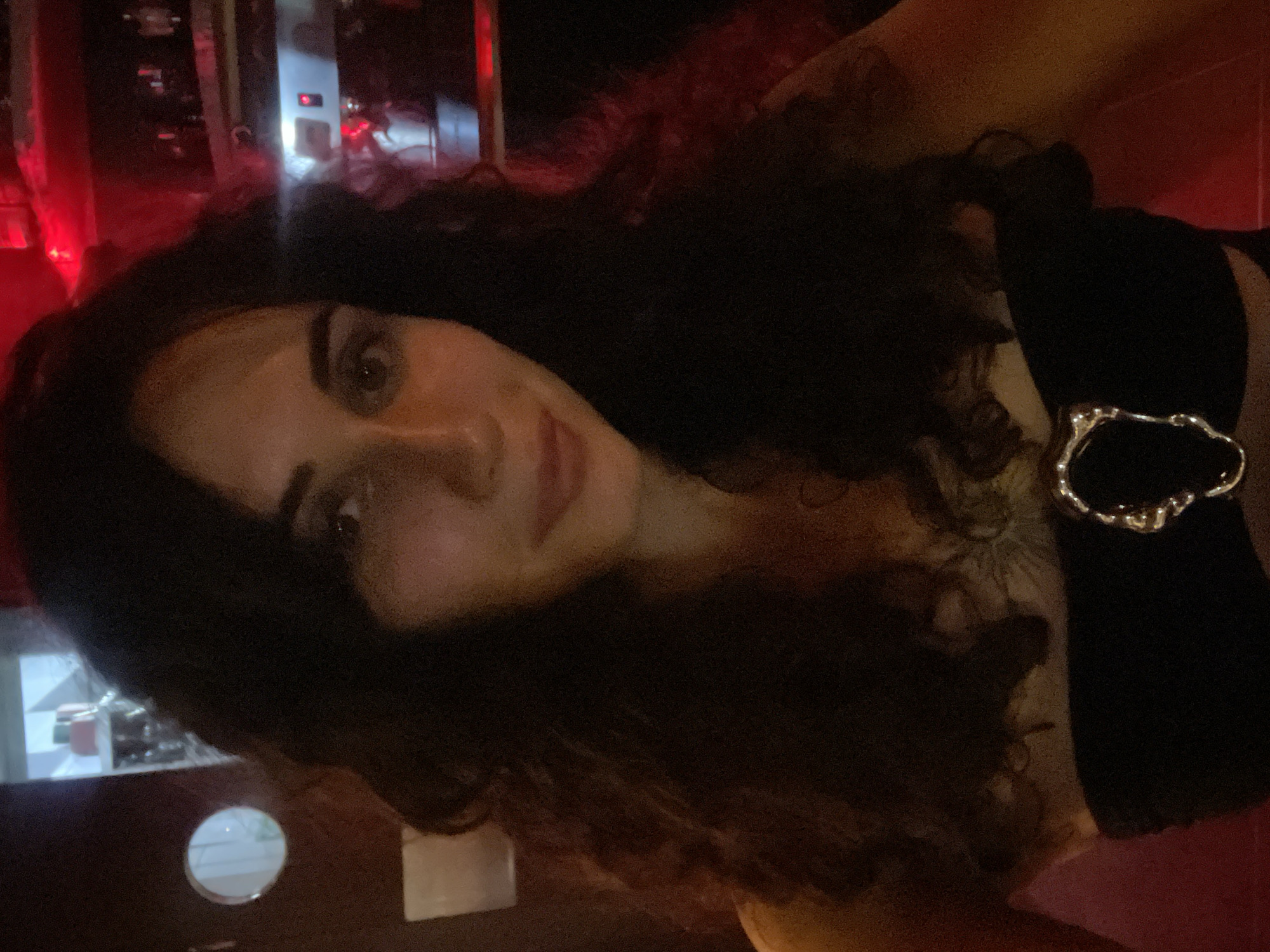 A young woman with dark, curly hair is pictured in a dimly lit indoor setting. She is wearing a strapless black top adorned with a silver metallic buckle. The background appears to be a bar or lounge, with hints of red and blue lighting, shelves with bottles, and a coffee machine. The woman is looking directly at the camera, with a slight smile. The image quality is somewhat grainy due to the low light conditions.