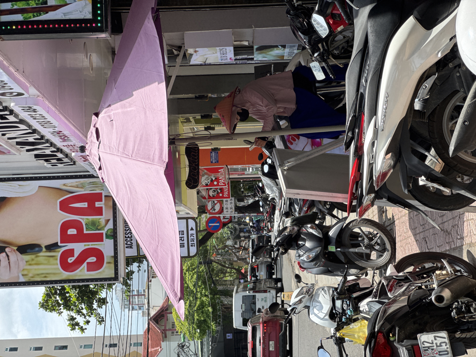 A street scene in Nha Trang, Vietnam, is depicted. In the foreground, several motorcycles are parked on the sidewalk. A pink umbrella shades a portion of the sidewalk. A person wearing a conical hat and a mask is seated under the umbrella, holding a phone. Behind the umbrella, a building with signage is visible. The sign reads "RELAX AND" and "SPA." Further down the street, traffic can be seen, including cars and other motorcycles. The environment appears to be a commercial area with buildings and trees lining the street.