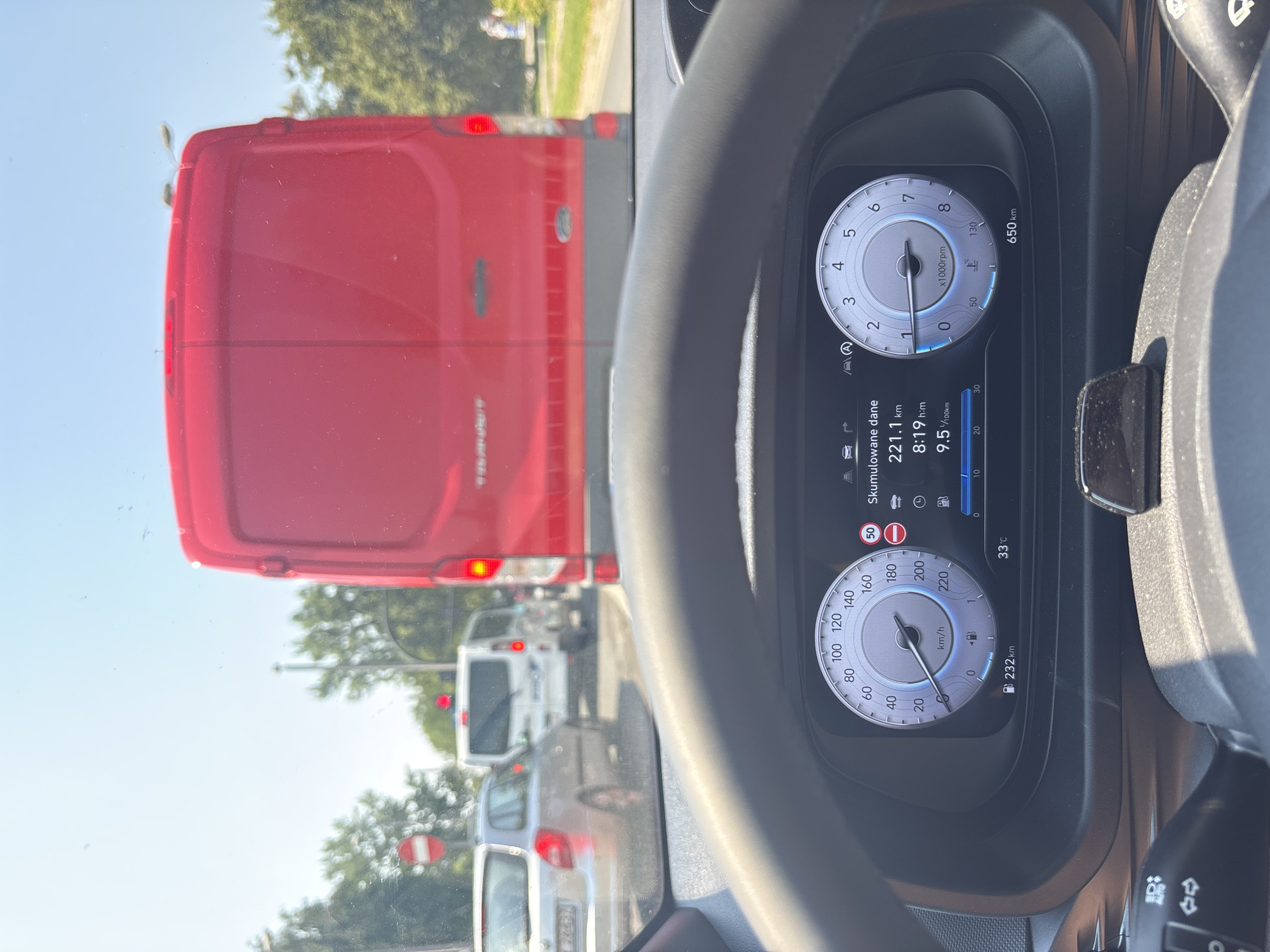 This is a photograph taken from the driver's perspective inside a vehicle, likely in Krakow, Poland, showing a traffic jam on a sunny day. The dashboard displays various information: the speedometer indicates the vehicle is traveling at 0 km/h, and the odometer shows a total of 232 km. A second digital display shows accumulated data: 221.1 km driven, 8:19 h time, and 9.5 l/100km fuel consumption. The temperature outside is 33°C. A tachometer is also visible, reading approximately 0 x1000rpm.
Outside the vehicle, traffic is at a standstill. In front of the car, a large red van is directly ahead. To the left of the red van, a silver hatchback is visible. Further ahead, a white van and other vehicles are stopped. Traffic lights are red, as indicated by the illuminated red circles on traffic signals and a "no entry" sign on the left. Trees and buildings line the street, suggesting an urban or suburban setting. The sky is clear and blue, indicating good weather conditions. The overall scene depicts a common urban scenario of being stuck in traffic.