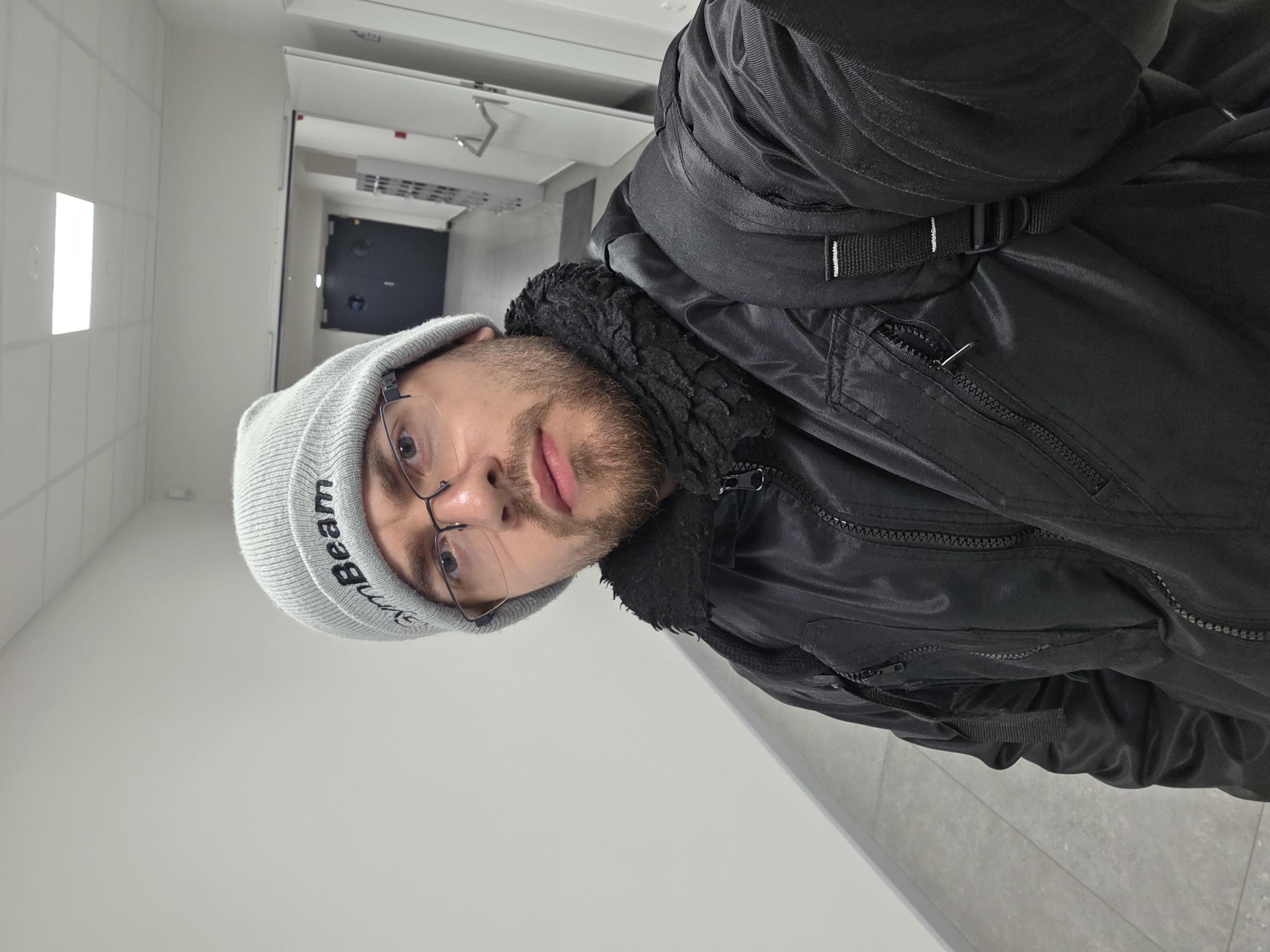 A person wearing a gray beanie with "Beam" written on it and glasses is wearing a black jacket with a fur-lined collar and a backpack. The person is standing in a hallway with white walls and ceiling tiles. There is a dark door at the end of the hallway. The location is in a building in Plovdiv, Bulgaria.