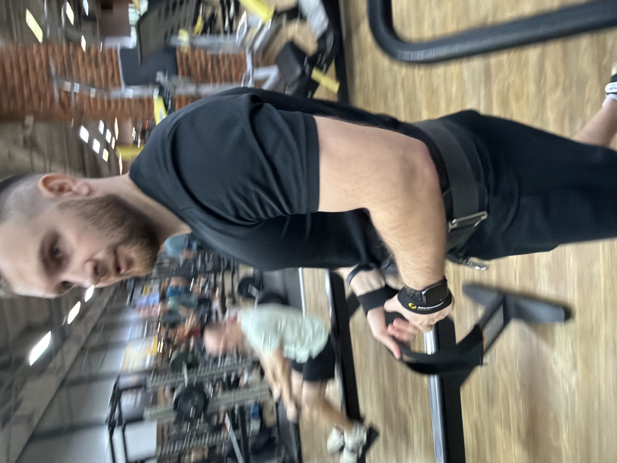 A man with a short beard and wearing a black t-shirt, black shorts, a weightlifting belt, and wrist straps with "ARUSHIHAN" visible on one, is working out at a gym. He is using a machine, possibly for rows or pull-ups, with black metal components. Other gym equipment and people are visible in the background, suggesting a populated workout environment. The location is in the city of Chaikovsky, Russia. The gym has a high ceiling with bright lights, and exposed brick is visible on one wall.