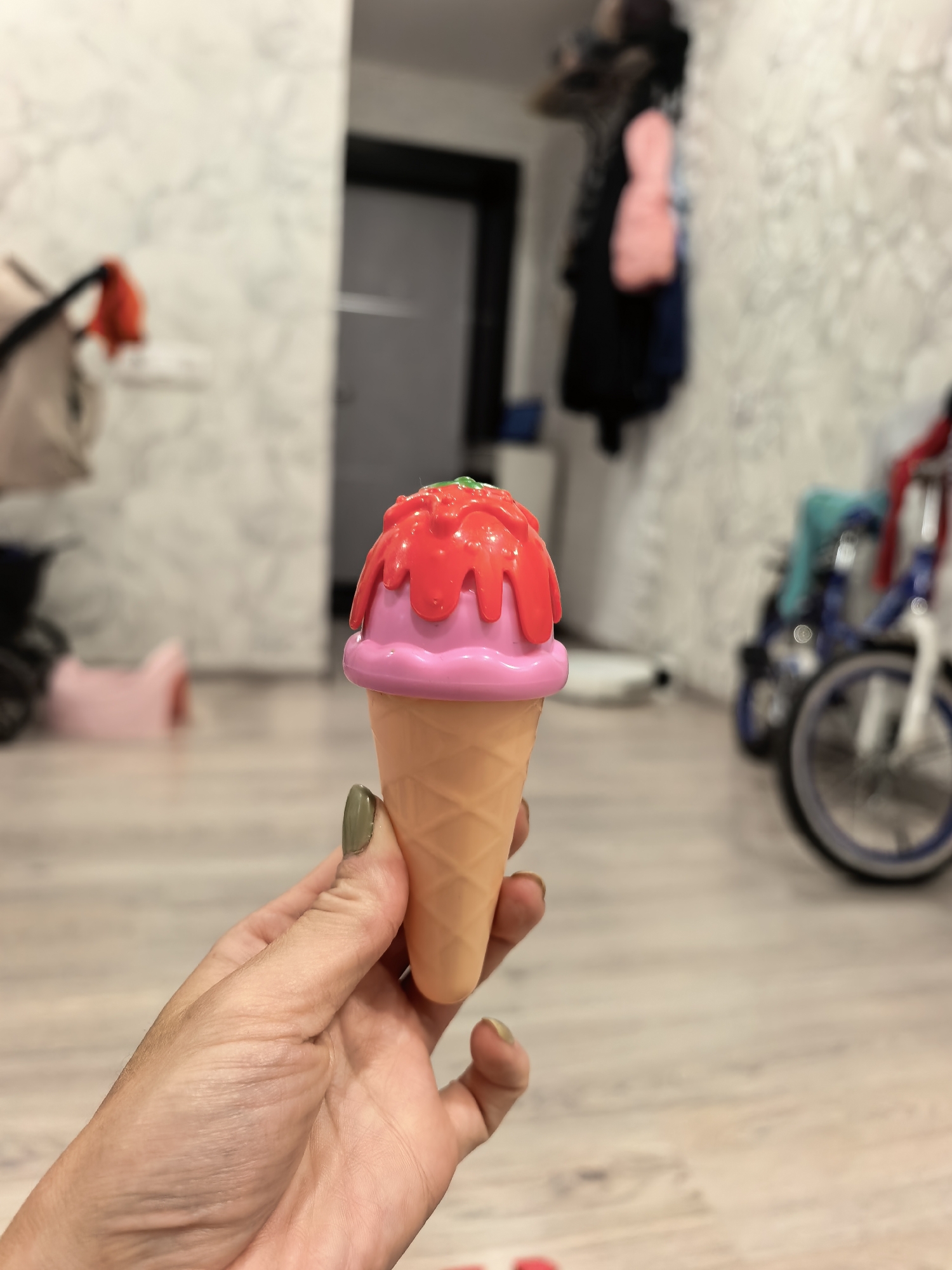 A person's hand with green nail polish holds a toy ice cream cone. The ice cream cone has a pink scoop and red topping. The background is blurred but shows a room with light-colored walls, a wooden floor, and a stroller and bicycle. The image is taken indoors.