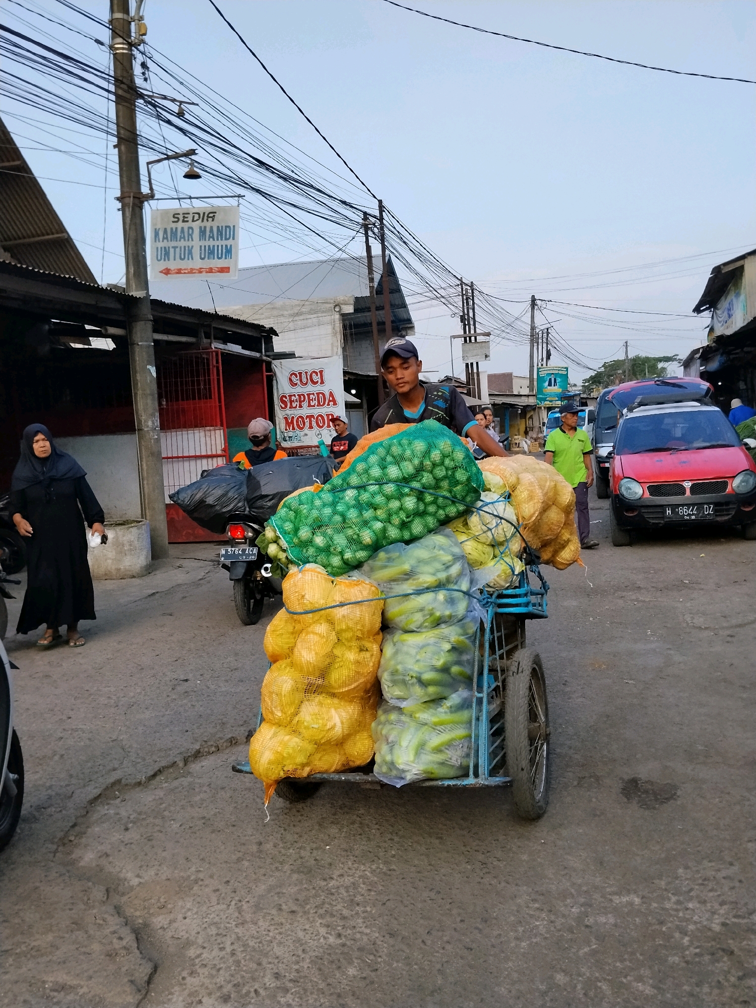 In Malang, Indonesia, a young man wearing a dark blue baseball cap and a dark blue and green t-shirt is pushing a cart loaded with produce. The cart is piled high with large net bags of green vegetables, yellow root vegetables, and what appear to be cucumbers. The man is on a street with cracked asphalt. To his left, a woman in a black hijab and black dress is walking away from the camera. Behind her, a motorcycle with license plate N 5764 ACA is parked. To the right of the cart, a red car with license plate H 8644 DZ is parked. Overhead, numerous electrical wires are strung across the sky. A sign on a pole to the left reads "SEDIA KAMAR MANDI UNTUK UMUM" (Toilets Available for Public Use) with an arrow pointing left. Another sign to the right, partially obscured, reads "CUCI SEPEDA MOTOR" (Motorcycle Wash). The buildings in the background appear to be shops.