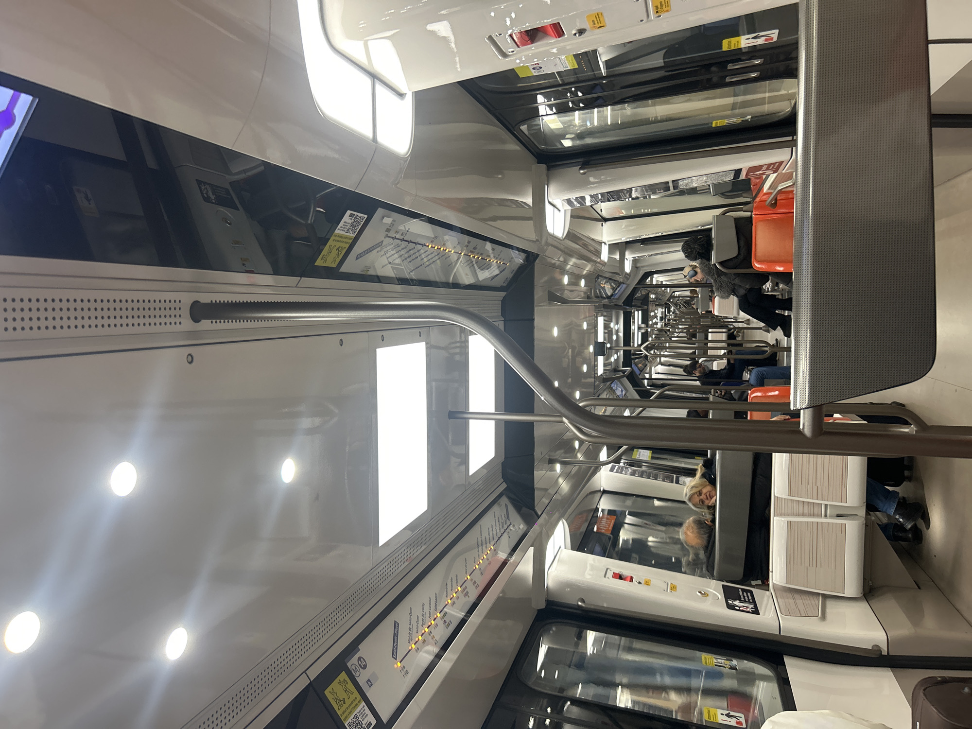 The image captures the interior of a train car, presumably a metro train in Paris, France, as indicated by the 'M 14' sign and station names such as "Saint-Denis-Pleyel" visible along the route map displayed above the windows. The carriage features a clean, modern design with white walls, windows along the top and sides, and a silver handrail that curves overhead. Passengers are seated throughout the car. The lighting is bright, casting reflections on the interior surfaces.