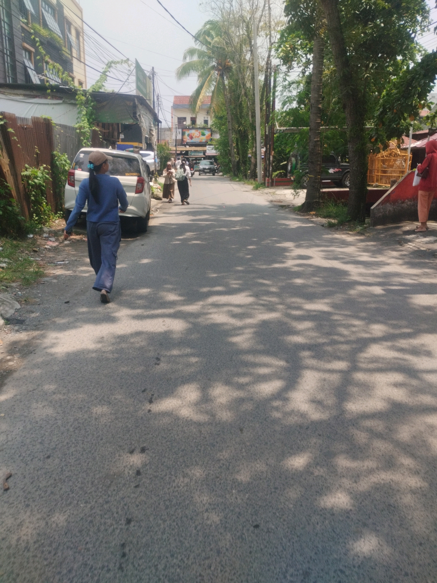 A person wearing a blue long-sleeved shirt, blue pants, and a tan cap walks away from the camera on a paved street. The person is on the left side of the street, moving towards the background. To the left of the person is a white car parked on the side of the road. A brown fence is visible behind the car. In the background, several other people are walking. Trees line the right side of the street, with their shadows cast across the pavement. Buildings and power lines are visible in the distance. The scene appears to be an outdoor urban environment, likely a residential or commercial street. The overall lighting suggests it is daytime with sunlight filtering through the trees. Based on the architecture and vegetation, the location is consistent with a street in the City of Medan, Indonesia.