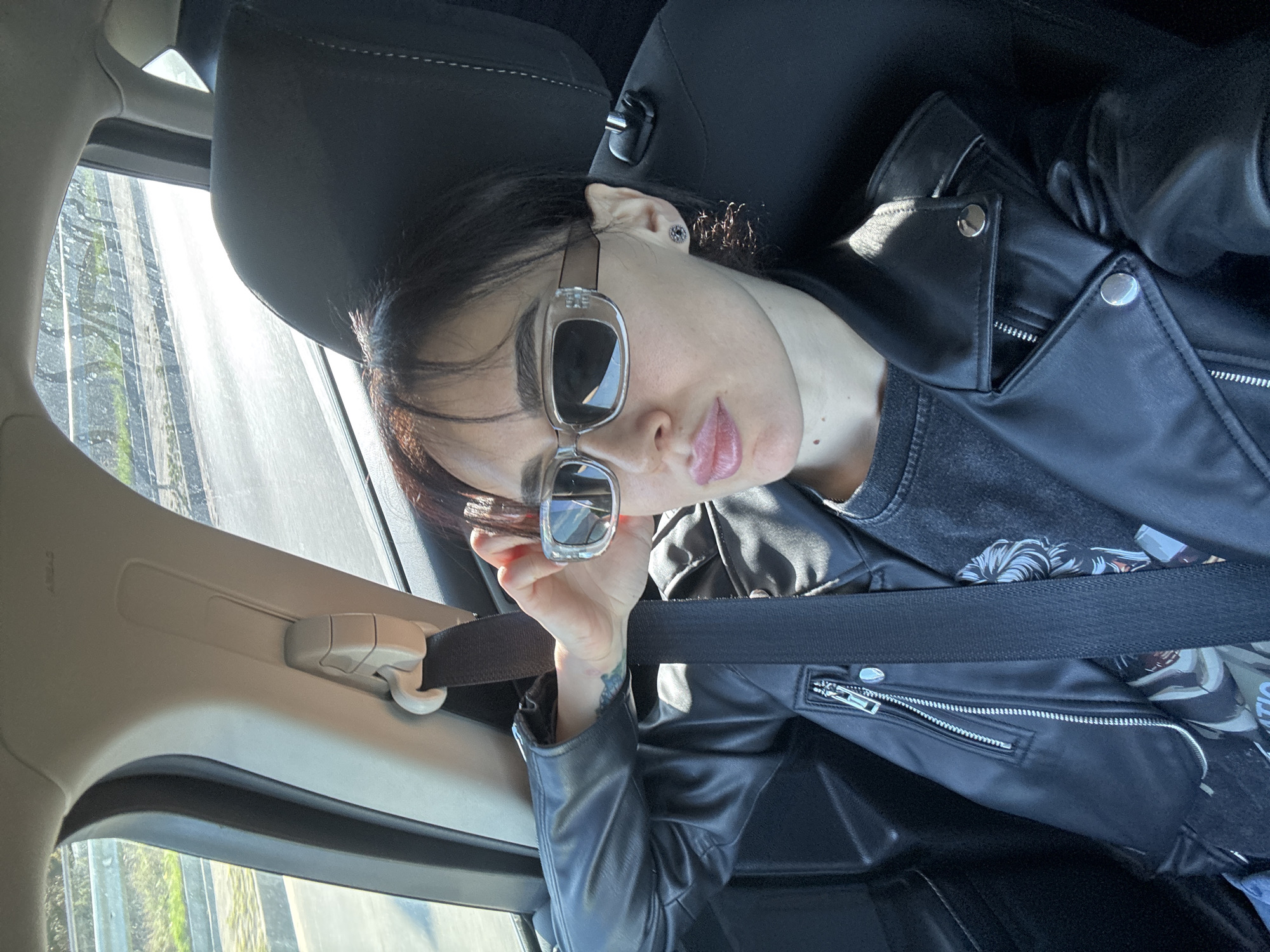 A medium shot of a person with dark hair and sunglasses, seemingly posing for a selfie in a car. The person is wearing a black leather jacket and a seatbelt. The background shows a blurred view from the car window, possibly indicating movement. The interior of the car is visible, with the person seated in the driver's seat. The location is challenging to determine definitively but based on the architectural details visible through the window, it may be possible that this scene is taking place in Tbilisi, Georgia.