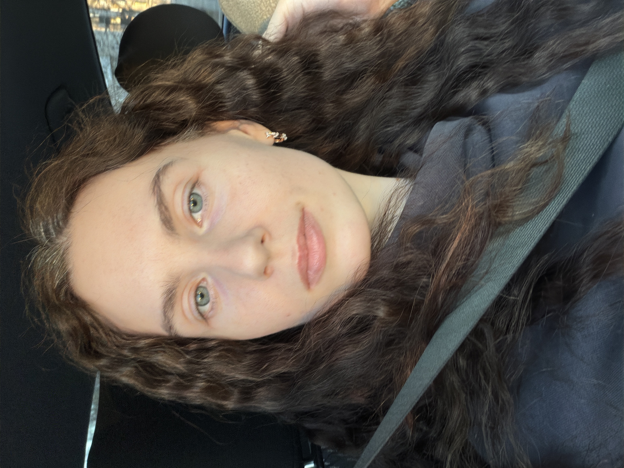 A young woman with fair skin and long, curly brown hair is seen in a close-up, tilted portrait. She has light green eyes and full lips. She appears to be wearing a seatbelt in a car. A small earring is visible in her left ear. The background includes elements of the car interior, such as the seat and window. There is no evidence to suggest this photo was taken in Saint Petersburg, Russia.