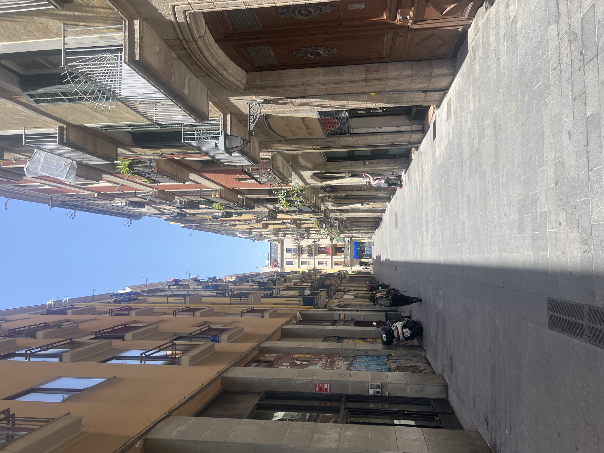 The image depicts a narrow street in Barcelona, Spain, lined with multi-story buildings featuring balconies and windows. The architecture suggests an older European city. The street is paved with stone slabs and is mostly empty, with sunlight casting long shadows.
On the left side of the street, a person with dark hair and a backpack walks away from the camera. Several motorcycles are parked along the curb. The buildings on this side are painted in warm, earthy tones, with some featuring graffiti.
On the right side of the street, a person in a pink shirt and dark pants is leaning against a building, possibly cleaning or inspecting it. This building has large wooden doors and arched doorways. Balconies with iron railings are visible on upper floors.
In the distance, the street continues, and more buildings can be seen, with a clear blue sky overhead. The overall impression is a quiet, sunlit street in a European city.