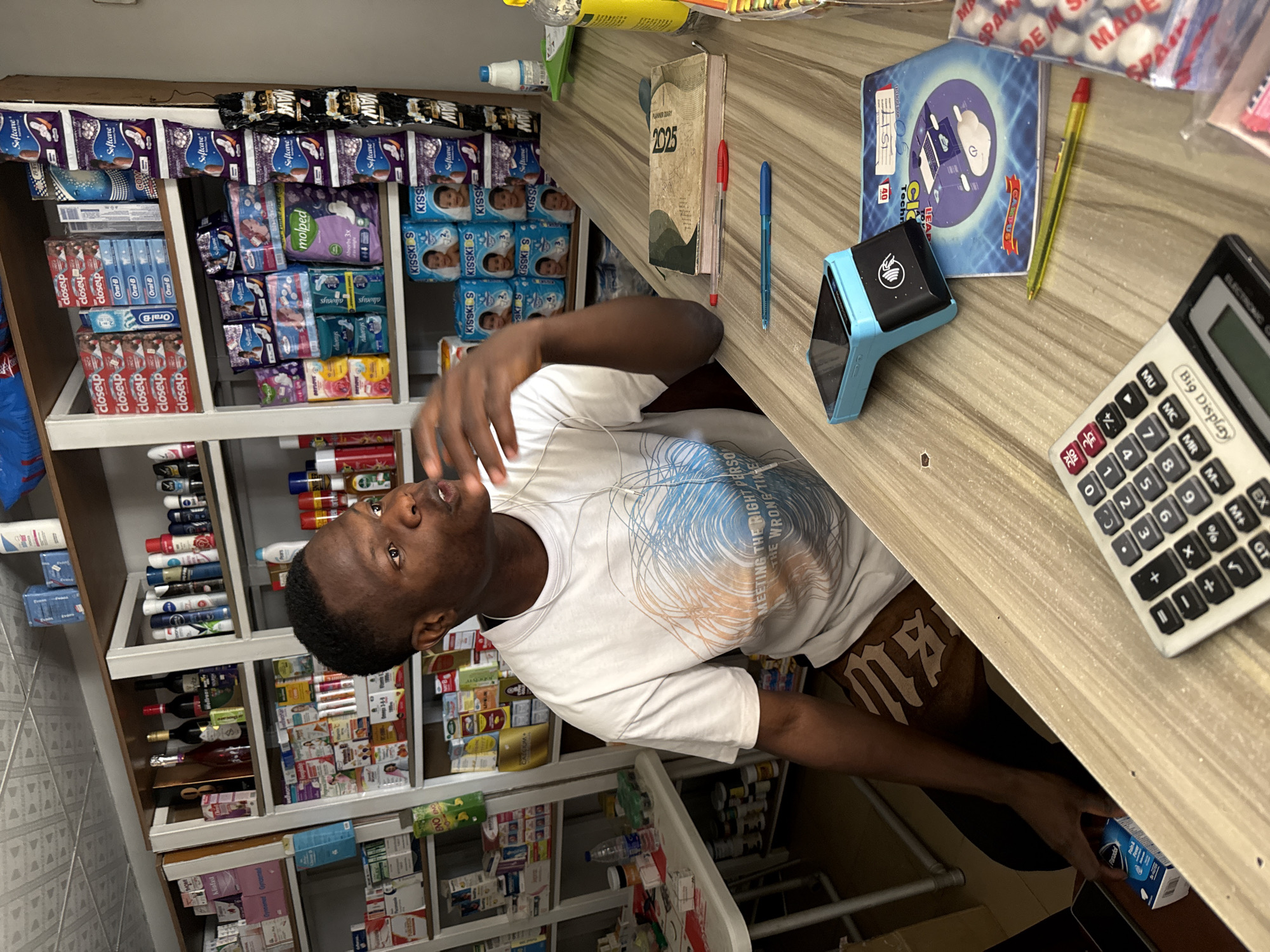 This media file shows a young Black man in a white t-shirt standing in a small convenience store or pharmacy. The store is stocked with various products, including sanitary pads, toothpaste, beverages, and other packaged goods, arranged on shelves. On the counter, there is a calculator, a card reader, a notebook, and pens. The man appears to be interacting with the items on the counter. The store is brightly lit, suggesting it might be daytime. The products on display include brands like "Softcare," "closeup," "Oral-B," and "always," suggesting the location is likely in Nigeria, possibly Abuja given the provided context. The overall scene depicts a typical retail environment.