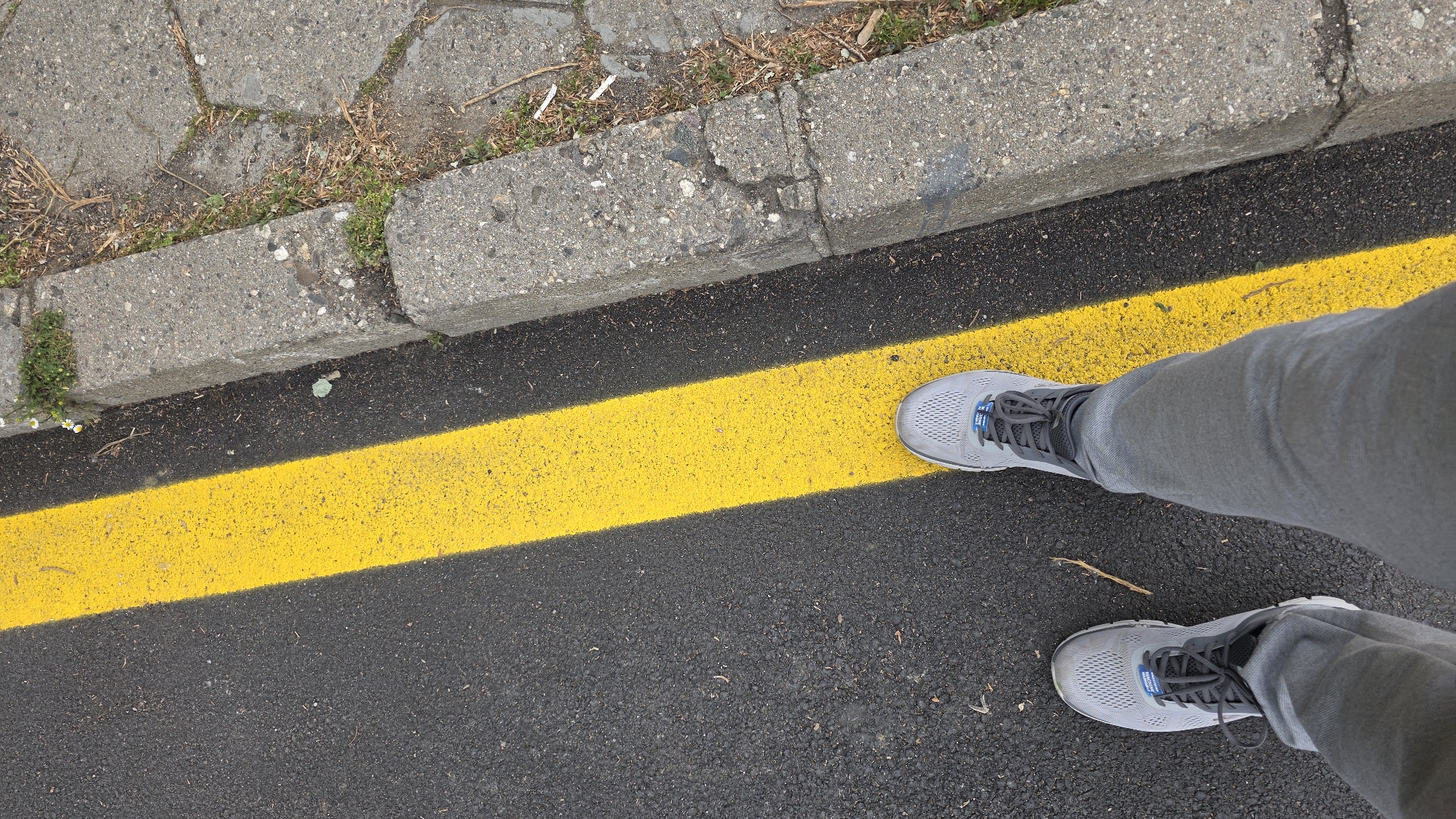The image is a first-person view of a person standing on an asphalt road in what appears to be an urban area. A solid yellow line is painted on the asphalt, and the person's grey running shoes and grey pants are visible. To the left of the yellow line, there is a concrete curb with some grass and weeds growing along it. The asphalt appears to be recently paved. The location is undetermined but presumed to be in Blagoevgrad, Bulgaria.