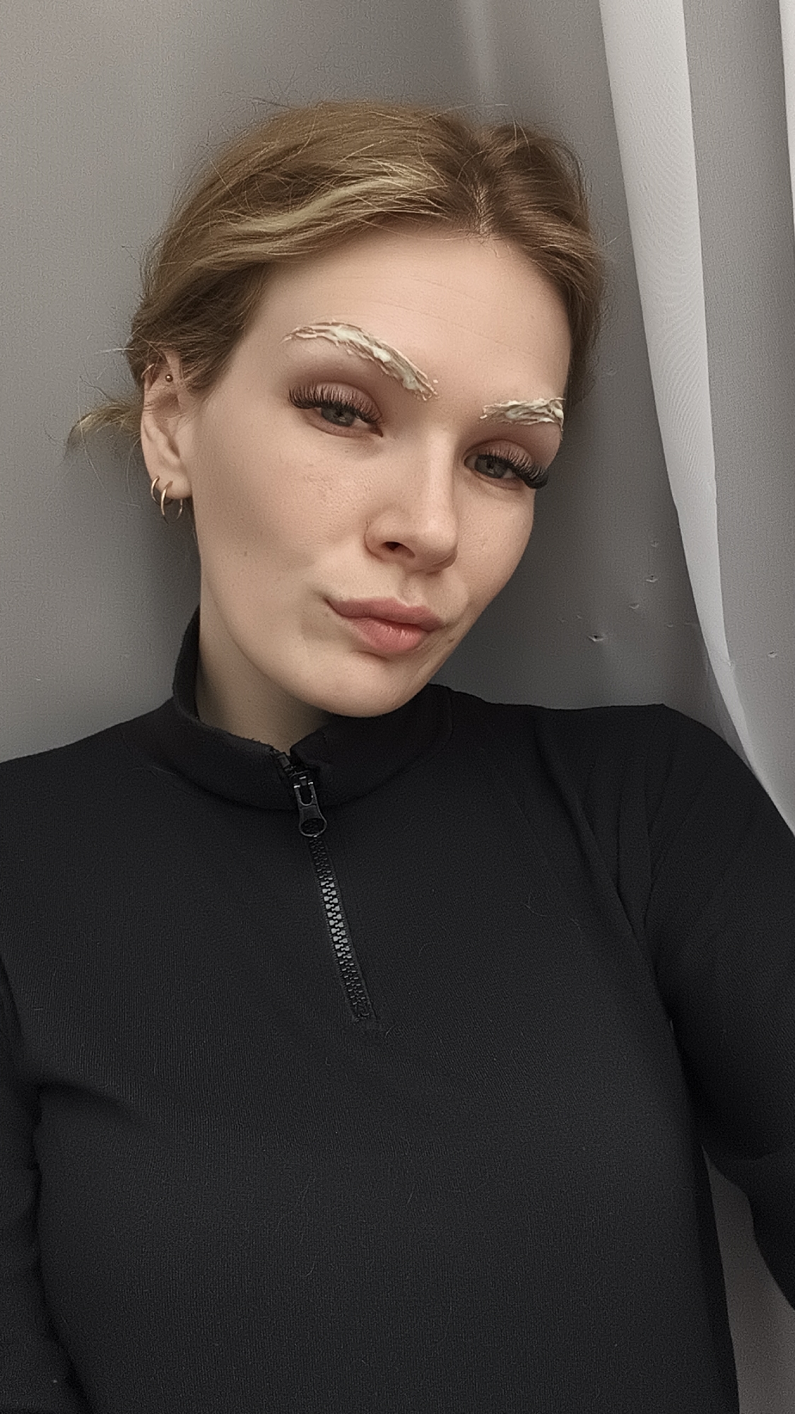 A young woman with fair skin and blonde hair is facing the camera, likely taking a selfie. She is wearing a black top with a partial zipper closure. Her eyebrows are covered in a white substance, possibly a cosmetic product or treatment. She has eyelashes, and is wearing earrings. The background consists of a gray wall on the left and a white curtain on the right. The location is specified as Русский Вожой, Russia, though there's no visual confirmation of this.