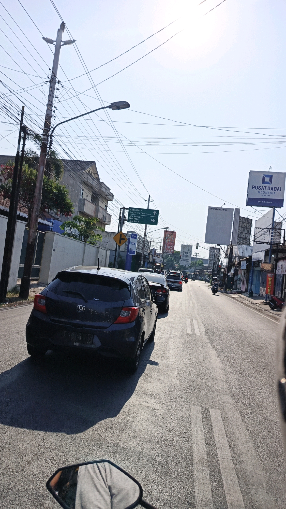 The image depicts a street scene in Surakarta, Indonesia, likely during daylight hours, with a clear sky. The perspective is from a vehicle, possibly a motorcycle, looking forward. The street is paved and appears to be a busy thoroughfare with multiple lanes of traffic. Several vehicles, including cars and motorcycles, are visible on the road. A dark-colored hatchback car is in the foreground on the left.
The urban environment includes buildings lining the street, some of which appear to be commercial establishments. Power poles with a dense network of wires dominate the upper portion of the image, along with a street lamp. Signage is present, including directional signs indicating "Kartosuro / Yogyakarta/Semarang" and "Sta. Purwosari / Stasiun Balai Kota," as well as advertisements and a sign for "PUSAT GADAI INDONESIA."
A person's leg and the mirror of a vehicle are visible in the lower part of the frame, indicating the viewpoint of the photographer. The overall impression is a typical street scene in an Indonesian city, characterized by moderate traffic and infrastructure elements.