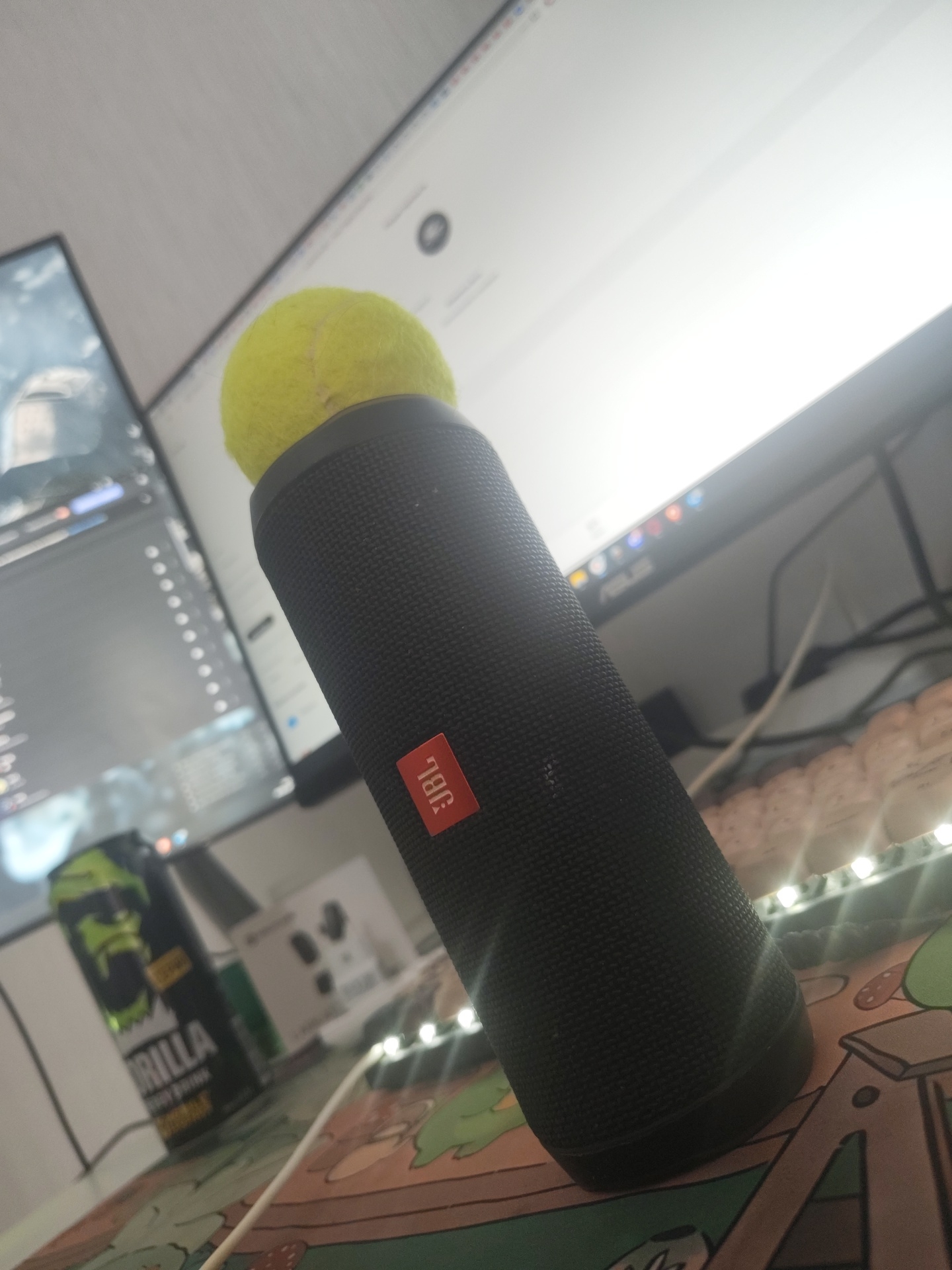 The image displays a close-up of a black cylindrical JBL speaker with a yellow tennis ball balanced precariously on top. The speaker has a prominent orange JBL logo on its side. In the background, a computer monitor is visible, showing a blurred image on its screen. To the left of the speaker, a green energy drink can with a gorilla logo is present. The objects are situated on a desk surface adorned with a cartoon-themed mat and string lights. The overall scene suggests a personal workspace or gaming setup. The image does not provide any information about the location in the city of Il’inskiy, Russia.