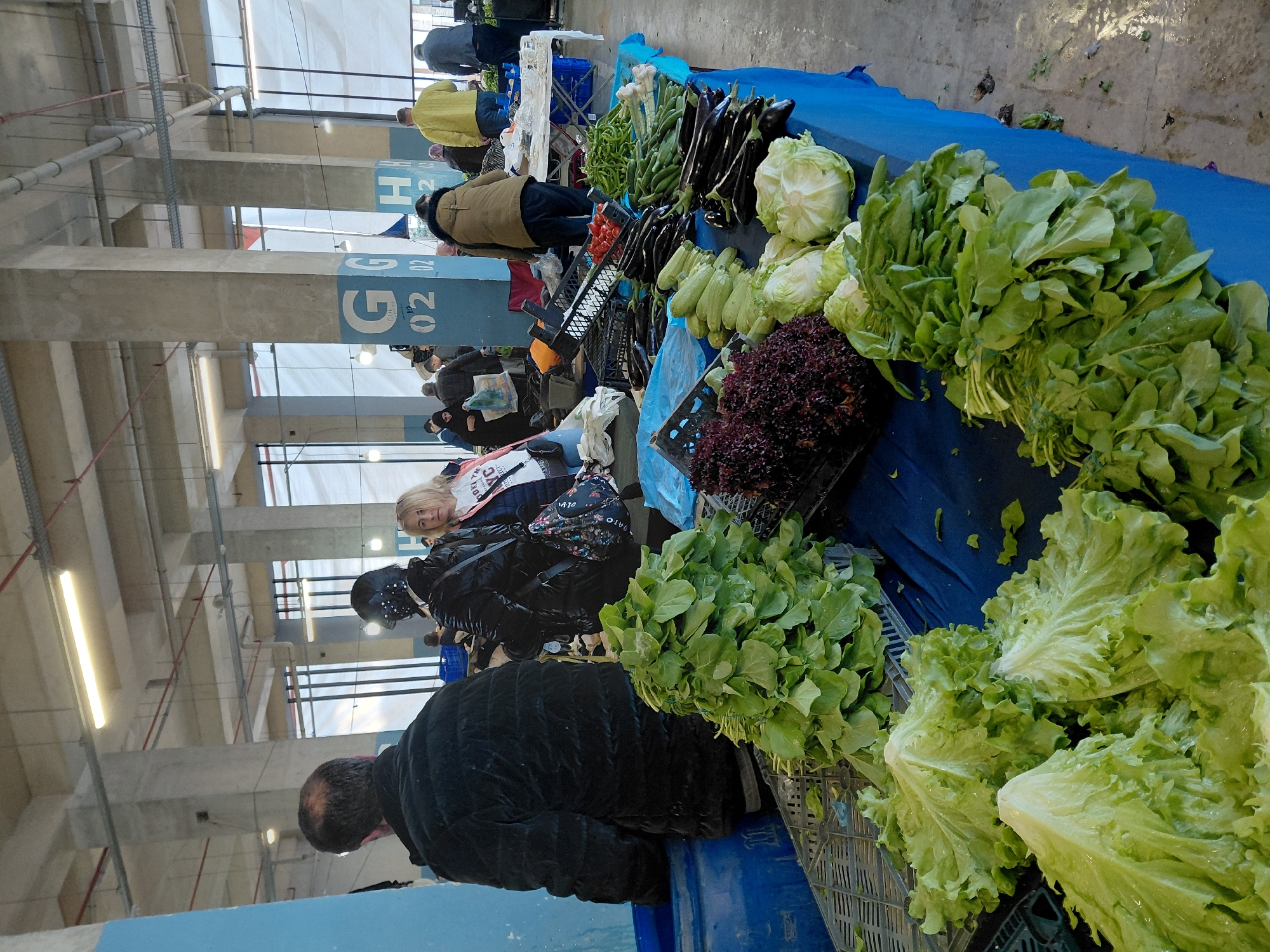 This image depicts a bustling indoor fresh produce market, likely a "pazar," in Üsküdar, Turkey. The scene is filled with vibrant displays of vegetables arranged on tables covered with blue tarpaulins and stored in black plastic crates.
In the foreground, prominent bunches of fresh green lettuce, including both loose leaf and compact heads, along with bundles of arugula and some red leaf lettuce, are showcased, many appearing freshly washed. Further into the market, a variety of produce is visible, including long purple eggplants, bright green peppers, light green zucchini, and clusters of red tomatoes.
Several people are engaged in the market activity. In the immediate foreground, a person in a black puffer jacket is seen from behind, leaning over a stall. In the mid-ground, a blonde woman, wearing a jacket and carrying a patterned shoulder bag, looks towards the camera. Other individuals, including shoppers and vendors dressed in warm clothing, are present throughout the background, browsing or tending to their stalls. A man in a yellow jacket is visible further back.
The market is housed within a large, covered structure featuring substantial concrete pillars and an exposed ceiling equipped with fluorescent lighting. A notable detail is a blue sign on one of the concrete pillars displaying "G 02 02", which likely indicates an aisle or section number within the market. The overall impression is of a lively local market offering fresh, seasonal produce.