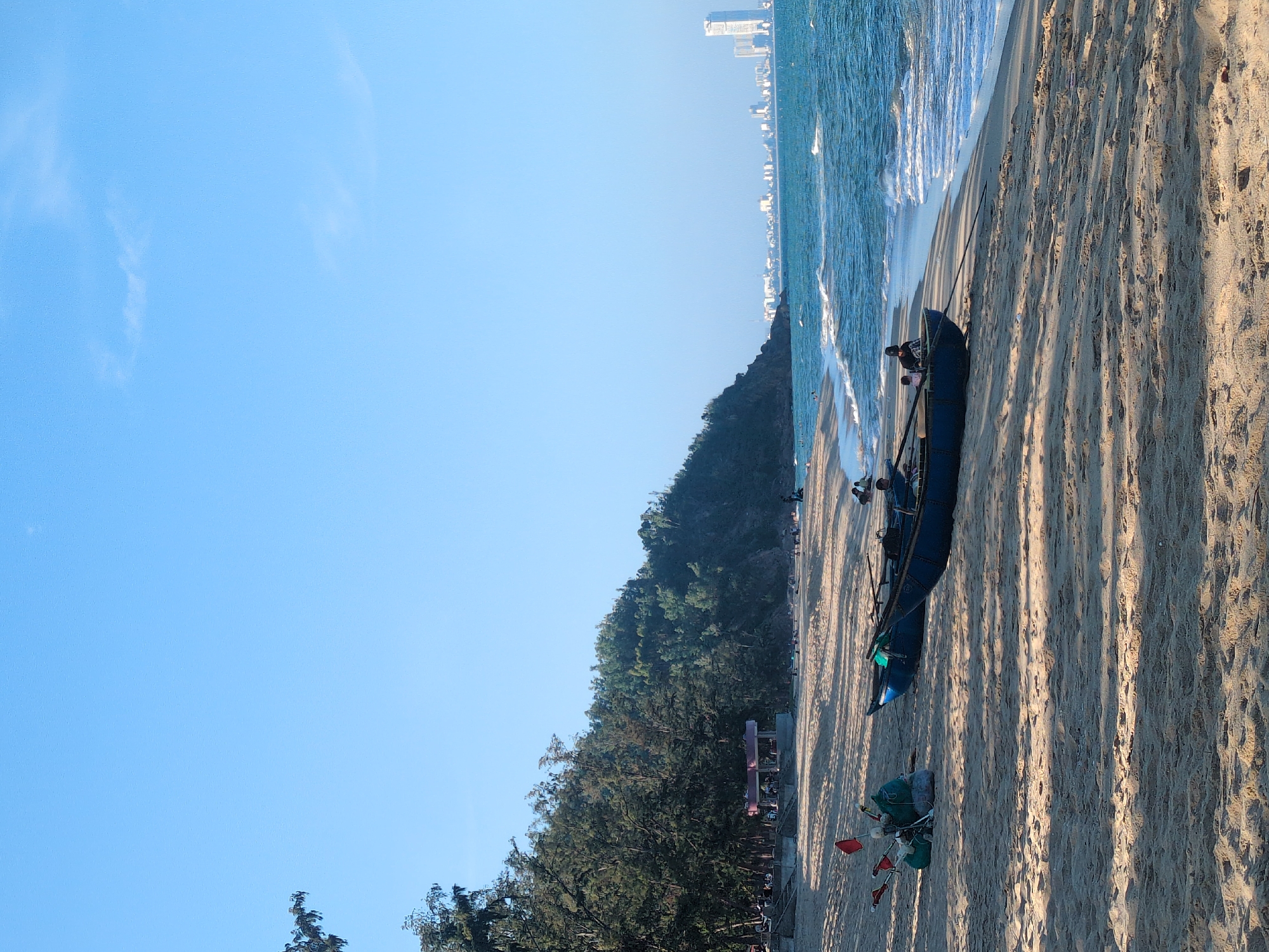 The image captures a beachfront scene in South Quy Nhon Ward, Vietnam. A fishing boat rests on the sandy beach, close to the shoreline where waves are breaking. To the left, a forested area provides a natural backdrop, while in the distance, a skyline of tall buildings is visible. The sky is clear and blue. There appear to be some people near the fishing boat. The overall impression is a serene blend of coastal life and urban presence.