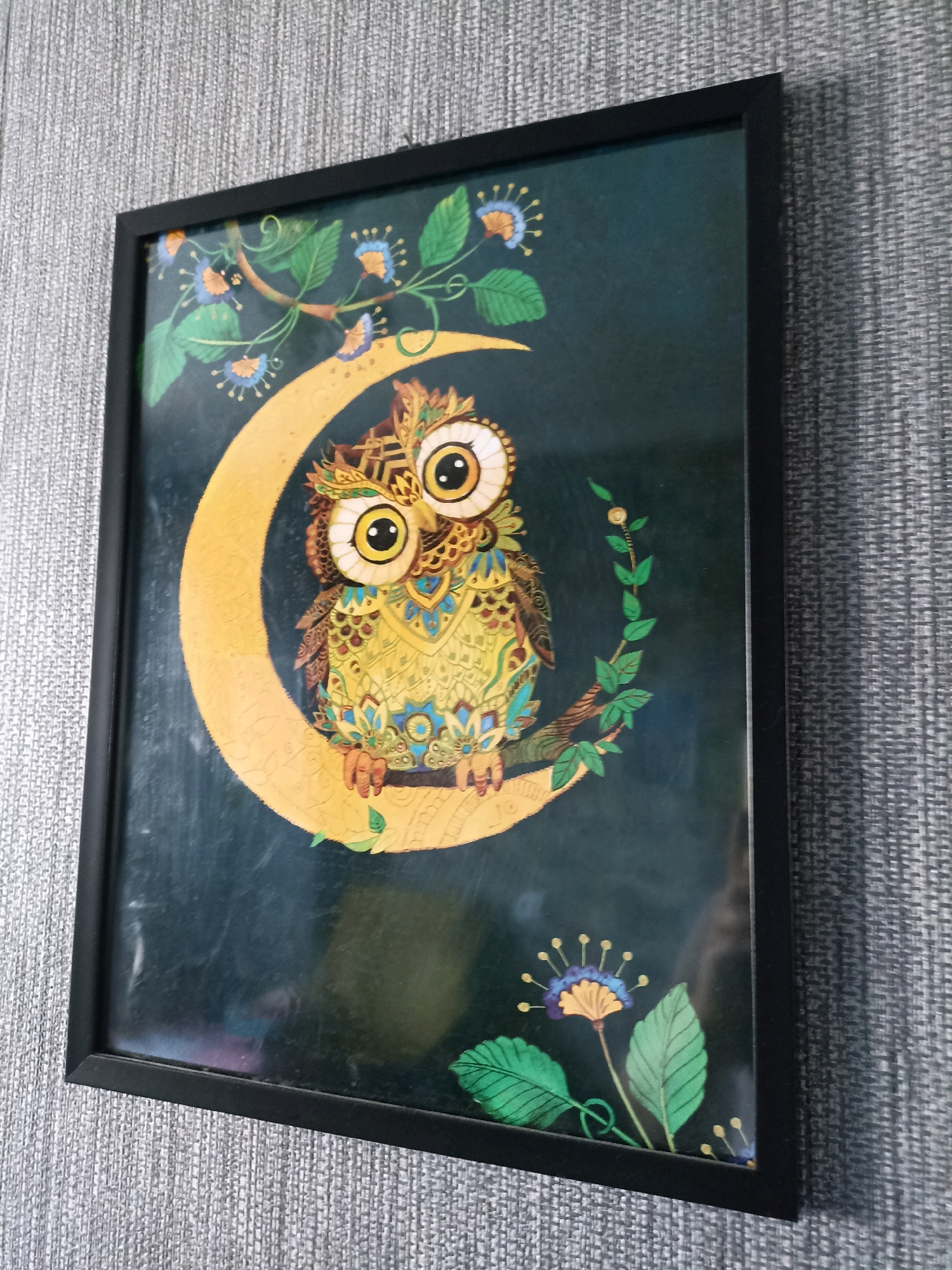 A framed picture of an owl sitting on a crescent moon is hanging on a textured gray wall. The owl is colorful, with large eyes and intricate patterns on its feathers. It is surrounded by stylized plants and flowers. The picture appears to be a piece of art or a print. The environment suggests an indoor setting, likely a home or office. The location of Novosibirsk, Russia is not discernible from the image itself.