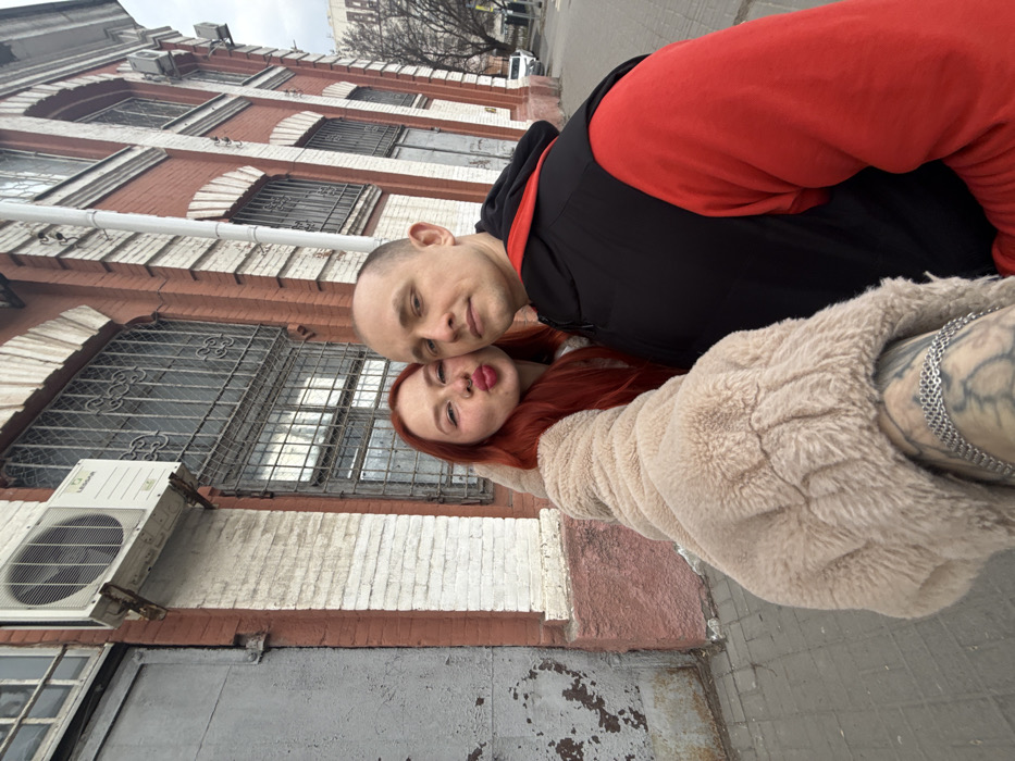 An outdoor selfie captures two individuals, a female and a male, in an urban setting identified as Rostov-on-Don, Russia. The female, positioned closer to the camera on the left, has long, reddish-brown hair, light skin, a nose piercing, and is wearing a light-colored top under a beige faux fur jacket. Her visible right arm displays a tattoo and a silver chain bracelet. She is looking towards the camera with puckered lips. The male, situated to her right and slightly behind, has a shaved head, light skin, and a neutral expression, dressed in a black sleeveless vest over a red long-sleeved shirt or hoodie.
The immediate background features a multi-story building constructed from red bricks with white decorative elements, including pilasters, cornices, and arched window surrounds, characteristic of older architecture in the city. The windows are fitted with dark, ornate metal security grates. An external air conditioning unit, branded "LESSAR," is mounted on the building wall above the female, exhibiting signs of wear and rust. To the far left, a large, weathered grey metal door or gate with peeling paint is visible. The ground directly in front of the building is paved with light-colored slabs. In the distant background to the right, a street is visible with bare trees and parked vehicles, including a white sedan, under an overcast sky.