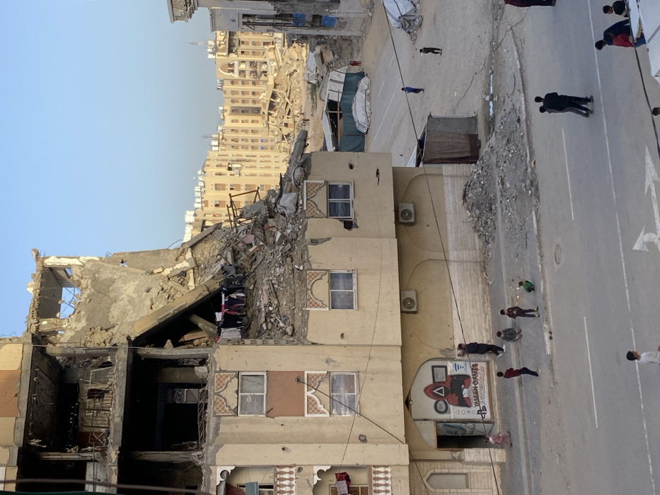 The image presents a scene of urban destruction, likely after a conflict or disaster, with signs of daily life continuing amidst the rubble. The view is from an elevated perspective looking down onto a street and damaged buildings.
On the left side of the frame, multiple multi-story buildings are severely damaged. The most prominent building has its upper floors completely collapsed, revealing exposed rebar, concrete slabs, and a large amount of debris and rubble. Despite the destruction, clothes are seen hanging to dry from a makeshift line in an exposed section of this building, indicating human presence or an attempt to salvage belongings. Adjacent to this, another building, while also showing signs of damage, has several intact floors. It features distinctive windows with decorative, arch-like designs above them and multiple air conditioning units mounted on its exterior walls.
In the foreground, on the lower part of one of the buildings facing the street, there is a large, colorful mural. The mural depicts a stylized figure in a red suit and black mask, holding a PlayStation controller, reminiscent of characters from "Squid Game." Below the character, the text "YASEEN GAMES" is clearly visible, along with the PlayStation logo and its iconic button symbols (triangle, circle, X, square).
The right side of the image features a paved street or road with white painted lines and a large white arrow pointing left, suggesting traffic flow. Along the edge of the street, particularly in the middle ground, are significant piles of rubble and debris, likely cleared from the damaged structures. In the distance, beyond the immediate scene of destruction, more urban buildings can be seen against a clear, light blue sky. Some of these distant buildings also appear to be damaged or partially collapsed.
Several people, including adults and children, are visible on the street and sidewalk. Some are walking, while others, particularly children, appear to be standing or playing near the roadside. In the middle distance, behind the damaged buildings and near the street, some temporary shelters or tents are set up, suggesting displacement or makeshift living arrangements.
The lighting suggests a clear day, possibly late morning or afternoon, with consistent illumination and soft shadows. The overall impression is one of stark devastation coexisting with human resilience and the continuation of daily routines in a difficult environment.