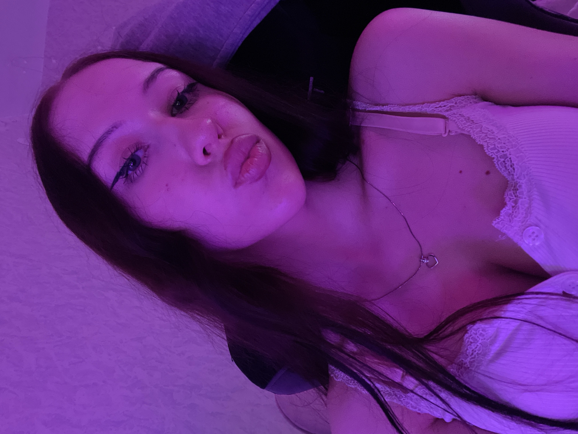 A young woman with long, dark hair is pictured in a close-up, indoor shot. She is wearing a light-colored camisole with lace trim and a delicate silver necklace with a heart-shaped pendant. Her makeup includes winged eyeliner. The lighting is predominantly purple, suggesting an ambient light source like a mood lamp or neon sign, which casts a pinkish hue on her skin and clothing. The background appears to be a textured wall. Her expression is a pout, with her lips pursed. The image does not contain any information that would allow for the determination of its location within Votkinsk, Russia.