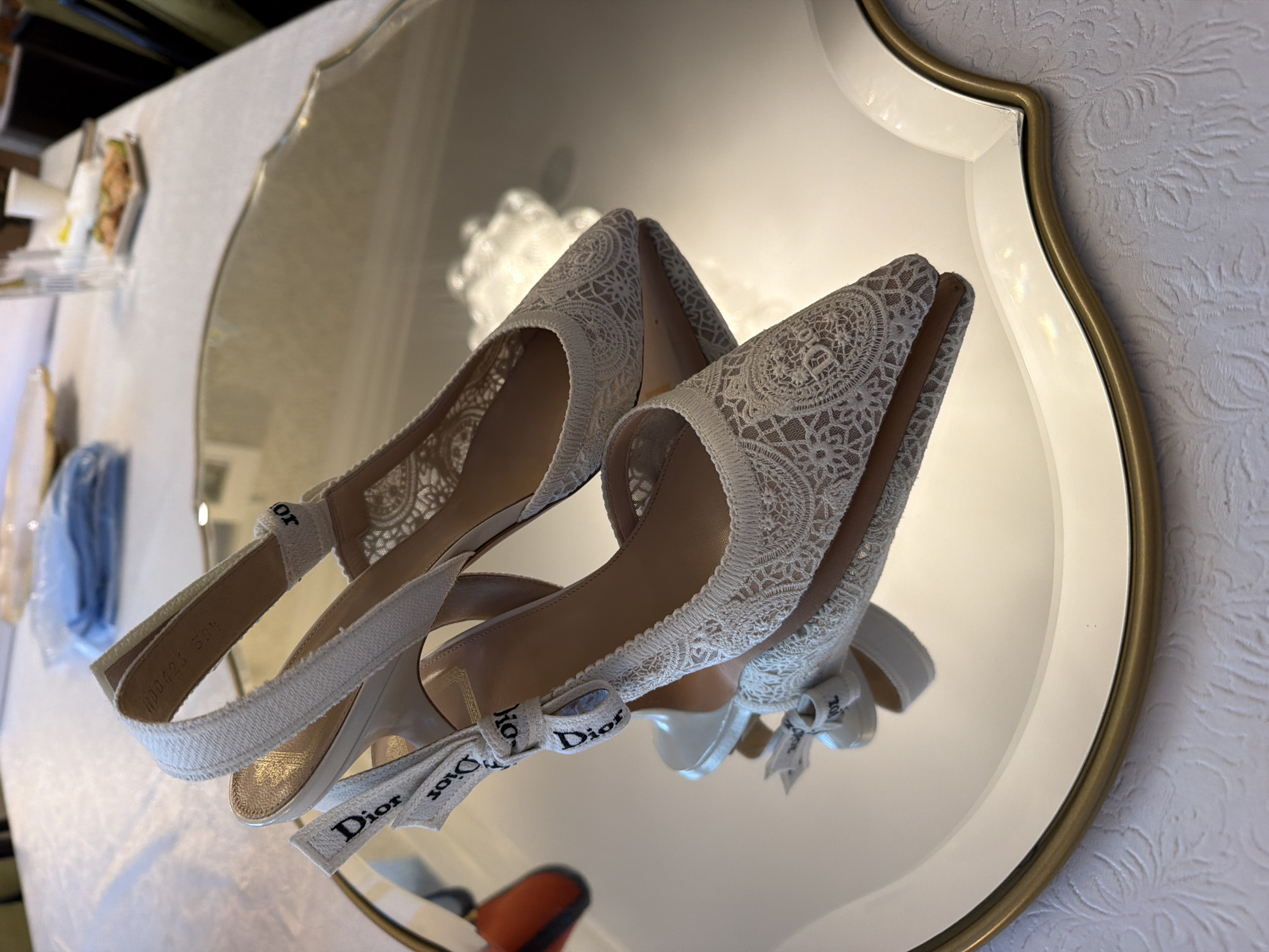 Two white lace Dior slingback heels are positioned on a mirrored surface with a scalloped edge, reflecting the shoes. The heels are adorned with a white ribbon featuring the Dior logo printed in black. The background shows a blurred image of a table with a white tablecloth, plates, and glasses, suggesting an indoor setting. The image appears to be taken in San Marino, United States of America.