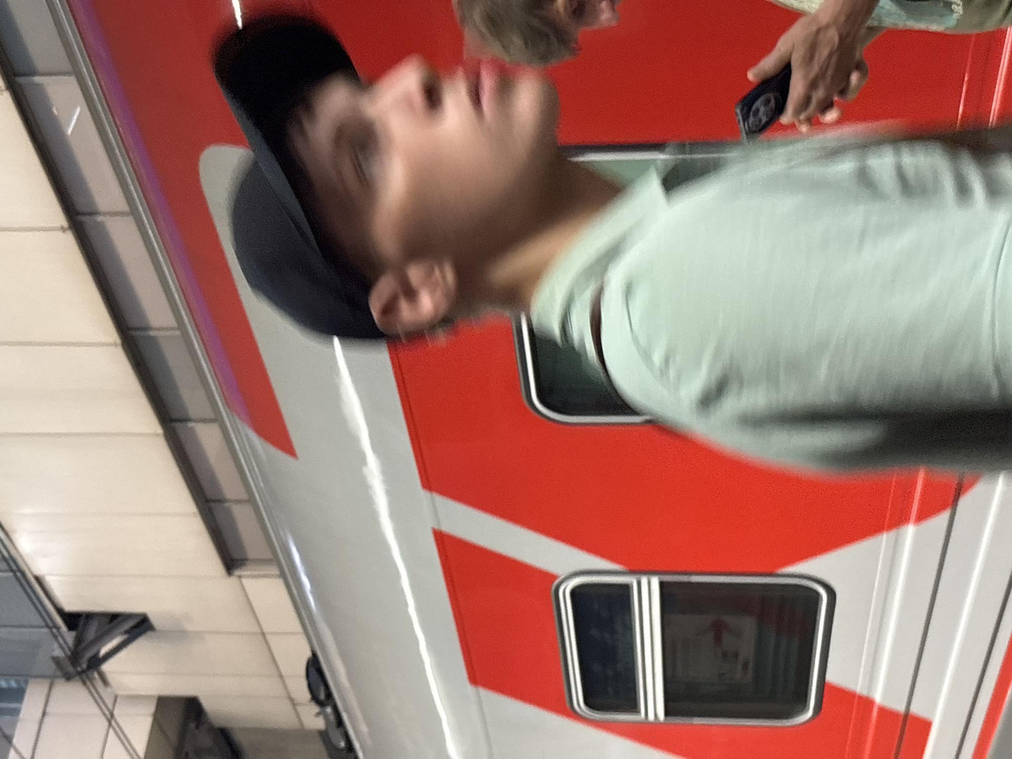 The image captures a young person wearing a black baseball cap and a light green collared shirt. The person is in profile view, facing towards the right of the frame, with their gaze directed slightly upward. The background consists of the side of a train with a prominent red and white pattern, suggestive of a Russian Railways train. The train has several windows, one of which is clearly visible in the foreground, displaying a red arrow pointing to the right. The scene appears to be at a train station, possibly within Moscow, Russia, given the context of Russian Railways. Another individual is partially visible to the right, holding a smartphone. The overall image has a shallow depth of field and exhibits motion blur, indicating movement or a fast shutter speed.