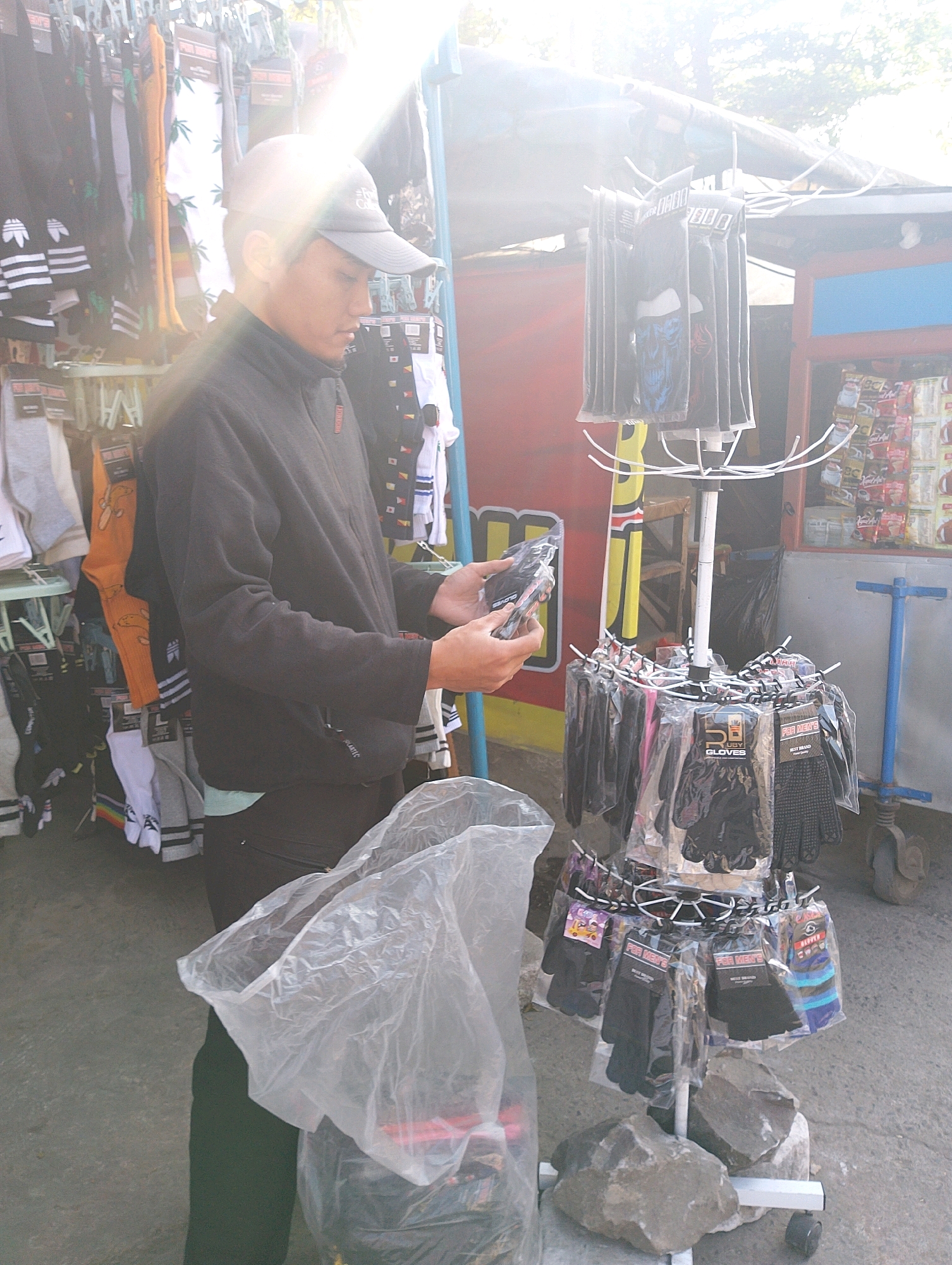 A young man wearing a black jacket and a grey baseball cap stands in front of a street stall in Lembang, Indonesia. The stall displays various items, including socks and gloves packaged in clear plastic. The man is holding a plastic bag and appears to be examining the merchandise. The background shows a busy street scene with other stalls and people. The lighting is bright, with a sun flare obscuring part of the image.