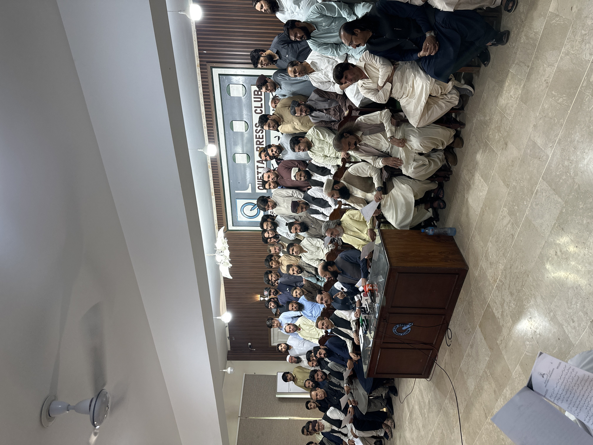 The image shows a group of men gathered indoors in what appears to be a press conference or meeting setting.
**People:** The group consists entirely of men, mostly dressed in traditional shalwar kameez. Their ages vary, and some wear head coverings.
**Subjects:** The central focus is a wooden podium with microphones. Some men stand or sit behind the podium.
**Objects:** A sign with the words "QUETTA PRESS CLUB" is visible in the background. Microphones, recording devices, papers, and water bottles are on the podium. A ceiling fan and light fixtures are visible.
**Actions:** Some men are speaking or presenting, while others appear to be listening.
**Interactions:** The interaction is primarily one of speakers addressing an audience.
**Environment:** The scene takes place in a room with light-colored walls and a tile floor. The press club sign and the presence of microphones suggest the location is the Quetta Press Club. The location is Quetta City Tehsil, Pakistan.