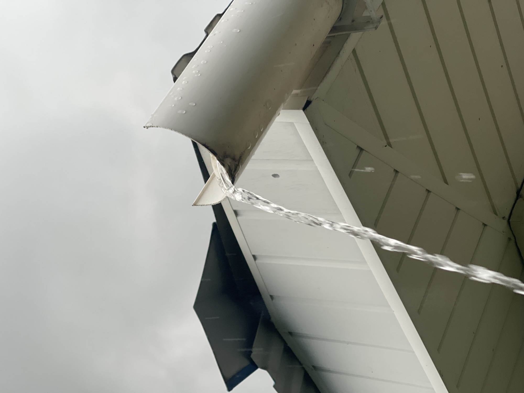 An outdoor, low-angle shot captures a white rain gutter on a building's eave, actively discharging a steady stream of water during an overcast day in the city of Unknown, Unknown. The sky above is uniformly light grey and cloudy. The gutter, positioned on the right side of the frame, is a half-round type, appearing to be made of white PVC or painted metal, with droplets of water clinging to its exterior surface. A notable dark, dirty stain is visible on the inner surface of the gutter where the water exits. The continuous stream of clear water cascades downwards from an opening in the gutter, angled towards the lower-left. Behind the gutter, dark, possibly black, roof shingles are partially visible, receiving light rainfall. Below the gutter and roofline is a white, horizontally paneled fascia or soffit structure. No individuals or other distinguishing objects are present in the image.