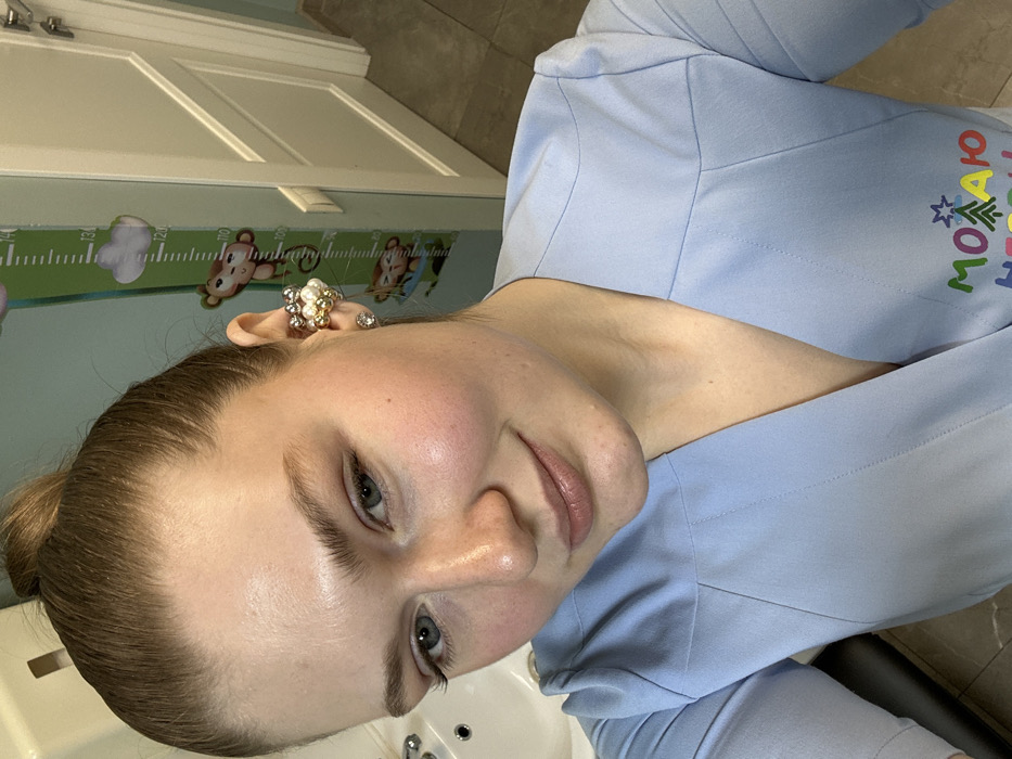 A fair-skinned person with light blue eyes and light brown hair pulled back in a bun is depicted at a close-up angle. The person is wearing elaborate pearl and gold earrings and a light blue top with colorful text. The background shows a white door with a growth chart adorned with cartoon animals. The person is positioned next to a white sink, which is partially visible. The image's orientation is rotated clockwise.