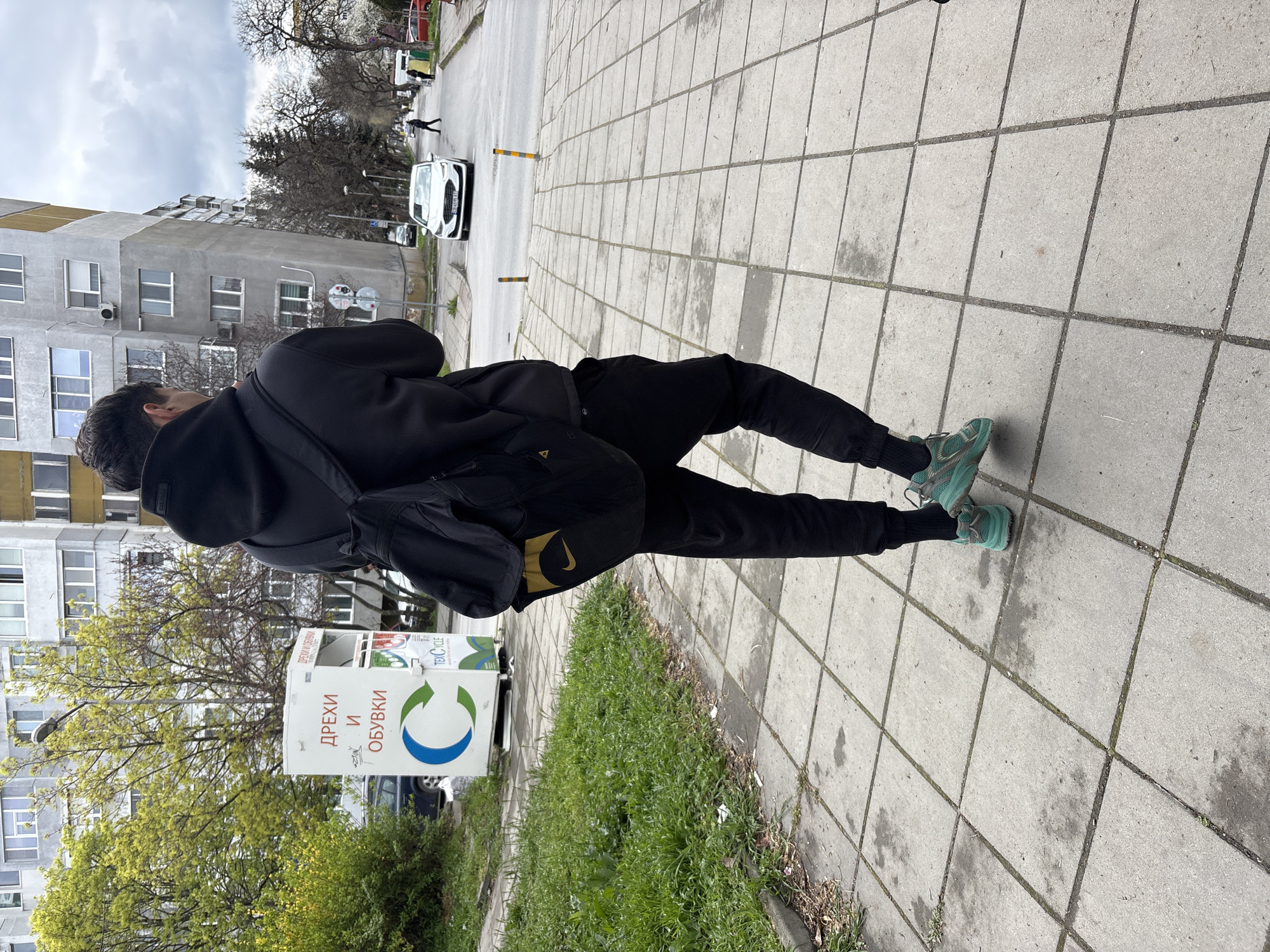 A man in black clothing and turquoise shoes is standing on a concrete tiled sidewalk. He is wearing a black hoodie, pants, and carrying a black Nike bag with a golden logo. The sidewalk is adjacent to a patch of green grass and a recycling bin labeled "ДРЕХИ И ОБУВКИ" (Clothes and Shoes) in Bulgarian. The background shows a gray apartment building and a street with a white car. The scene appears to be in Varna, Bulgaria, based on the Bulgarian text on the recycling bin. The sky is overcast.