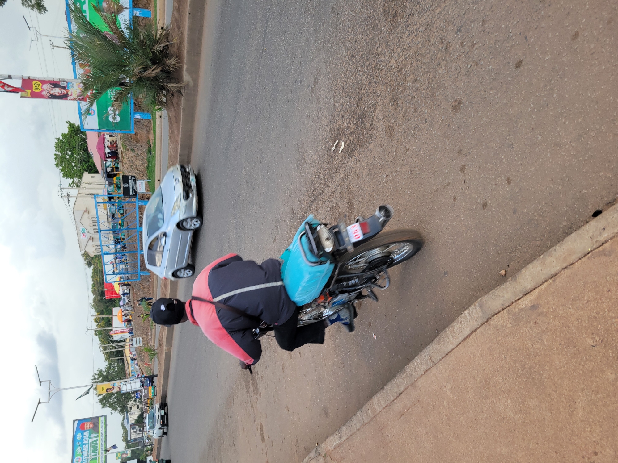 The image captures a street scene in Jos, Nigeria, with a motorcycle rider in the foreground. The rider, wearing a black cap, a red and blue jacket, and black trousers, is seen from behind, operating a motorcycle with a bright blue seat cover. The motorcycle has a visible license plate with red lettering and white numbers that appear to read "053." The motorcycle is on a paved road with a concrete sidewalk to the left.
In the background, various elements indicate an urban environment. There are several vehicles on the road, including a silver hatchback car and what appear to be rickshaws or tricycles. Billboards and signs are visible, some advertising political campaigns and businesses. One billboard features an image of a person with the text "NG. GOVT. AND DISTANCE APPLS, PLATEAU STATE." Another displays a smiling person with the text "Yes!". Buildings and trees line the street, and power lines are suspended overhead. The sky is overcast with clouds. The overall scene depicts a typical day on a busy road in Jos.
