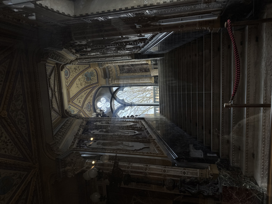 The scene depicts the interior of a grand staircase hall within the Doge's Palace (Palazzo Ducale) in Venice, Italy. The vantage point is at the base of a wide, upward-leading stone or marble staircase, which occupies the central and lower portions of the frame. The staircase features dark, polished marble balustrades and lighter-colored steps. A red velvet rope, secured by two brass stanchions, acts as a barrier across the bottom steps.
The walls flanking the staircase are ornately decorated. The lower sections are paneled with dark, veined marble. Above this, lighter panels feature rectangular artworks, possibly frescoes or paintings, depicting figures, framed by elaborate decorative borders. Columns and pilasters with intricate capitals are integrated into the wall design. A dark, ornate light fixture with multiple globe-shaped bulbs is visible on the left wall. The vaulted ceiling above is extensively decorated with intricate painted patterns in muted golds, blues, and creams, set within recessed panels and mouldings.
At the top of the staircase, a large, multi-paned Gothic-style window with pointed arches is prominently featured, emitting bright natural light that illuminates the upper part of the staircase and creates visible light rays within the shadowed interior. Through the window, the bare branches of trees are visible against a bright, possibly overcast, sky, along with a glimpse of an outdoor balustrade. The overall lighting is dim in the foreground and lower sections of the hall, contrasting with the brightness from the window. No individuals are present in the scene.