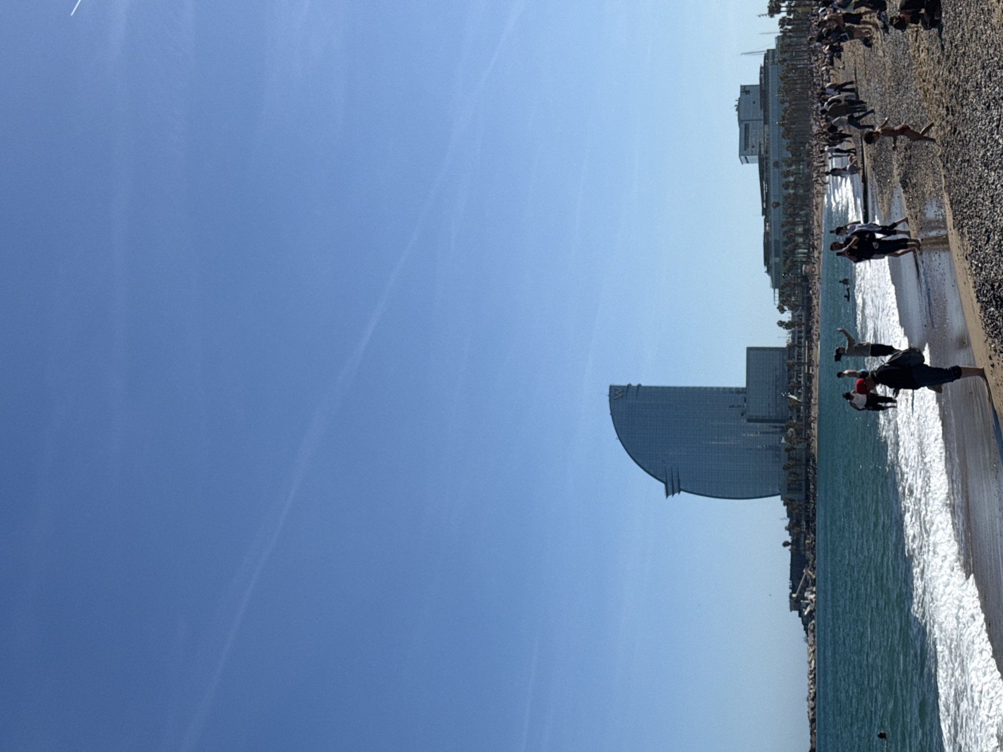 The image depicts a sunny day on Barceloneta Beach in Barcelona, Spain. The Hotel Arts Barcelona is prominently visible in the background. People are seen on the beach and in the water. The beach appears to be pebbled. The sky is clear blue.