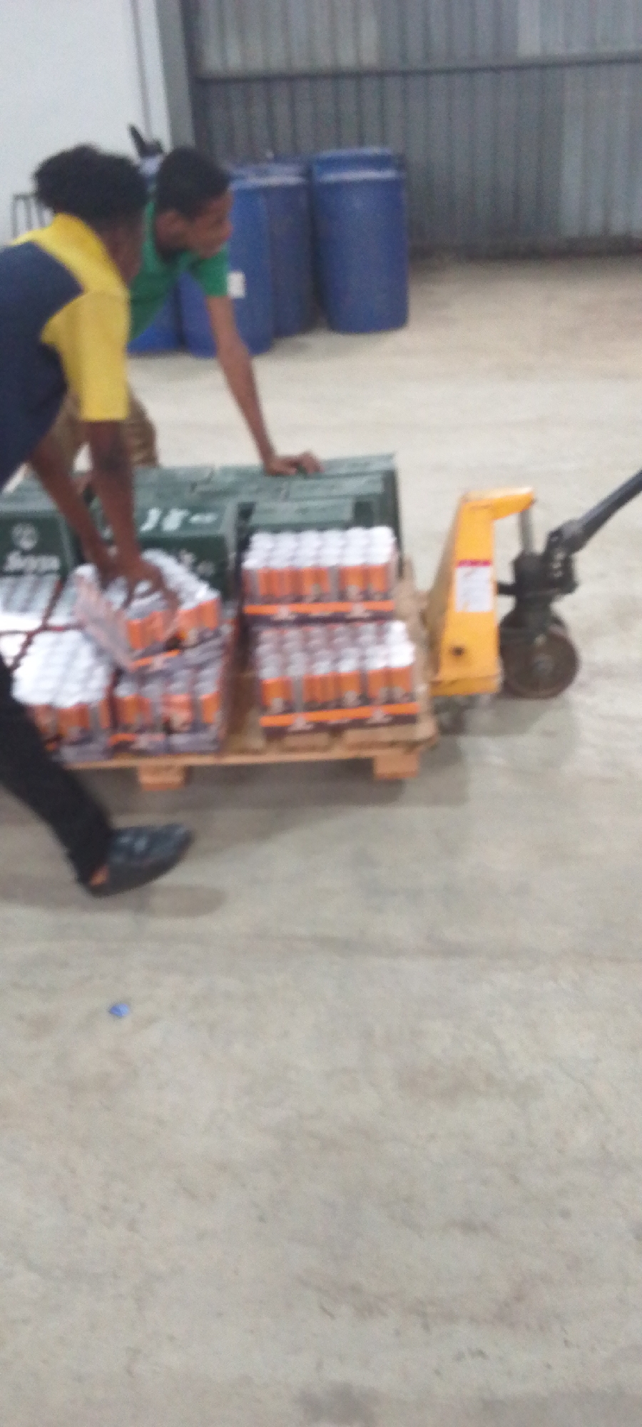 In an indoor warehouse setting, two individuals are observed transporting goods on a pallet jack. The individual on the left is wearing dark trousers and black footwear, a shirt with navy and yellow sections. The person on the right is wearing a green t-shirt. They are jointly maneuvering a pallet jack laden with multiple packages of canned beverages and a green crate. The pallet jack is primarily yellow with black components. The warehouse background includes blue barrels and a metal wall.