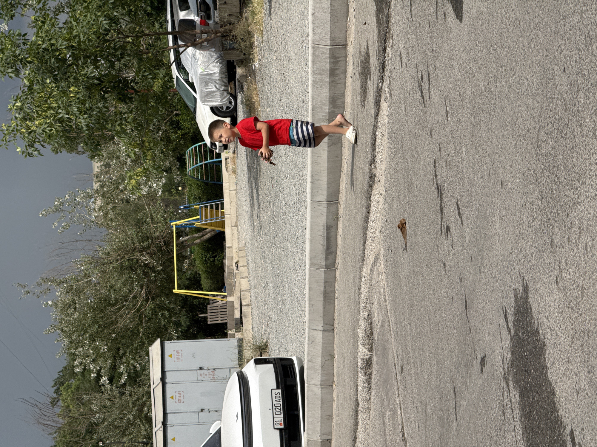 In an outdoor scene, a young boy in a red t-shirt and striped shorts stands on a paved surface. He is barefoot except for white slides on his feet and is holding a small object in his hands. He looks towards the left of the frame. In the background, a white car with a license plate reading "G1 02G ADS KG" is parked. Beyond the car, a playground with yellow and blue structures is visible, along with trees and a gray industrial building. The sky is overcast and dark. The ground in the foreground appears wet in patches.