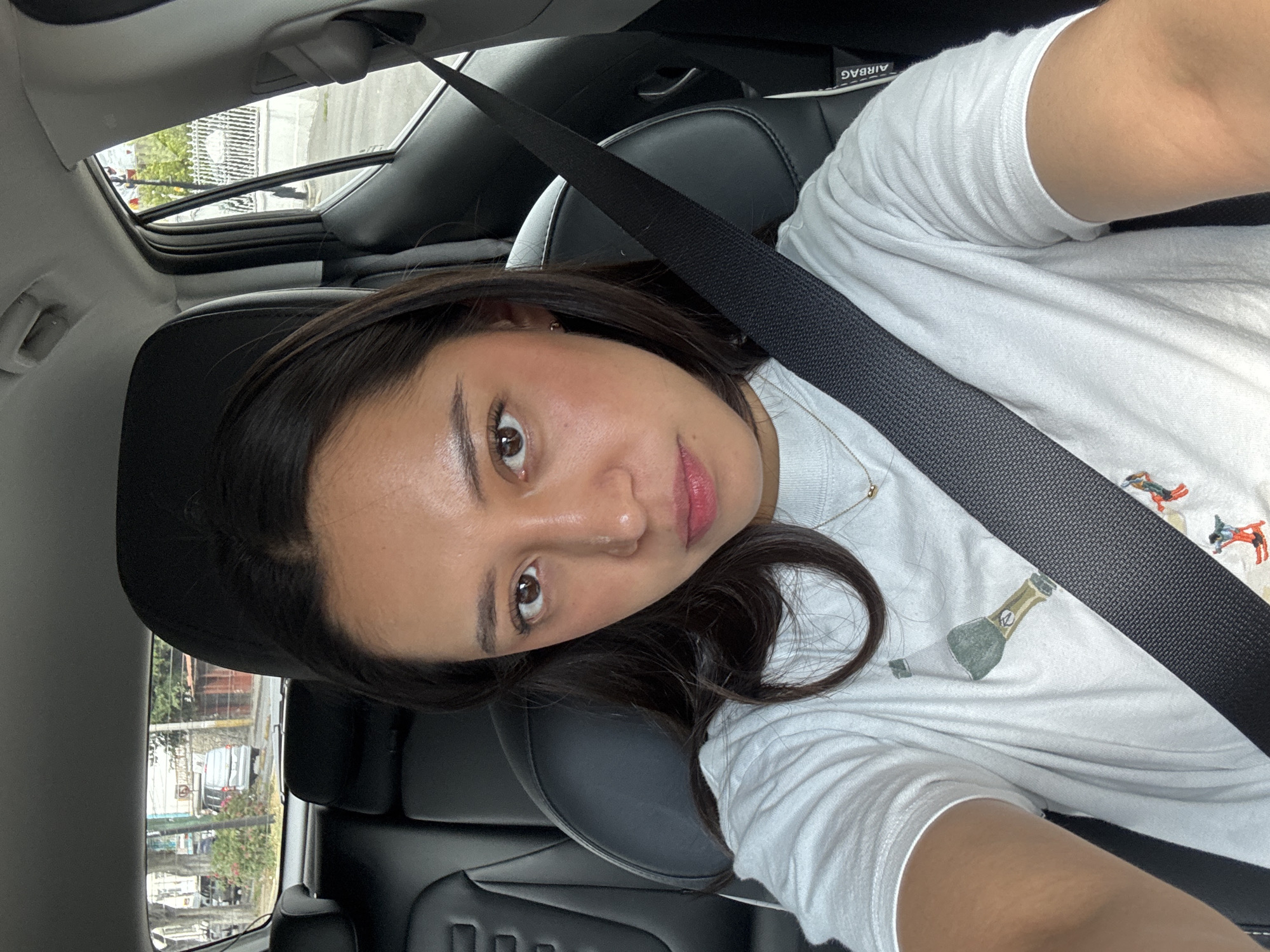A woman with shoulder-length dark brown hair is sitting in the driver's seat of a vehicle. She is wearing a white t-shirt with an image of champagne bottles. A seatbelt is fastened across her chest. The vehicle has a black interior and a sunroof. Outside the window are buildings and trees typical of an urban environment. Based on the architecture and general environment, the location is likely in the city of Puebla, Mexico.