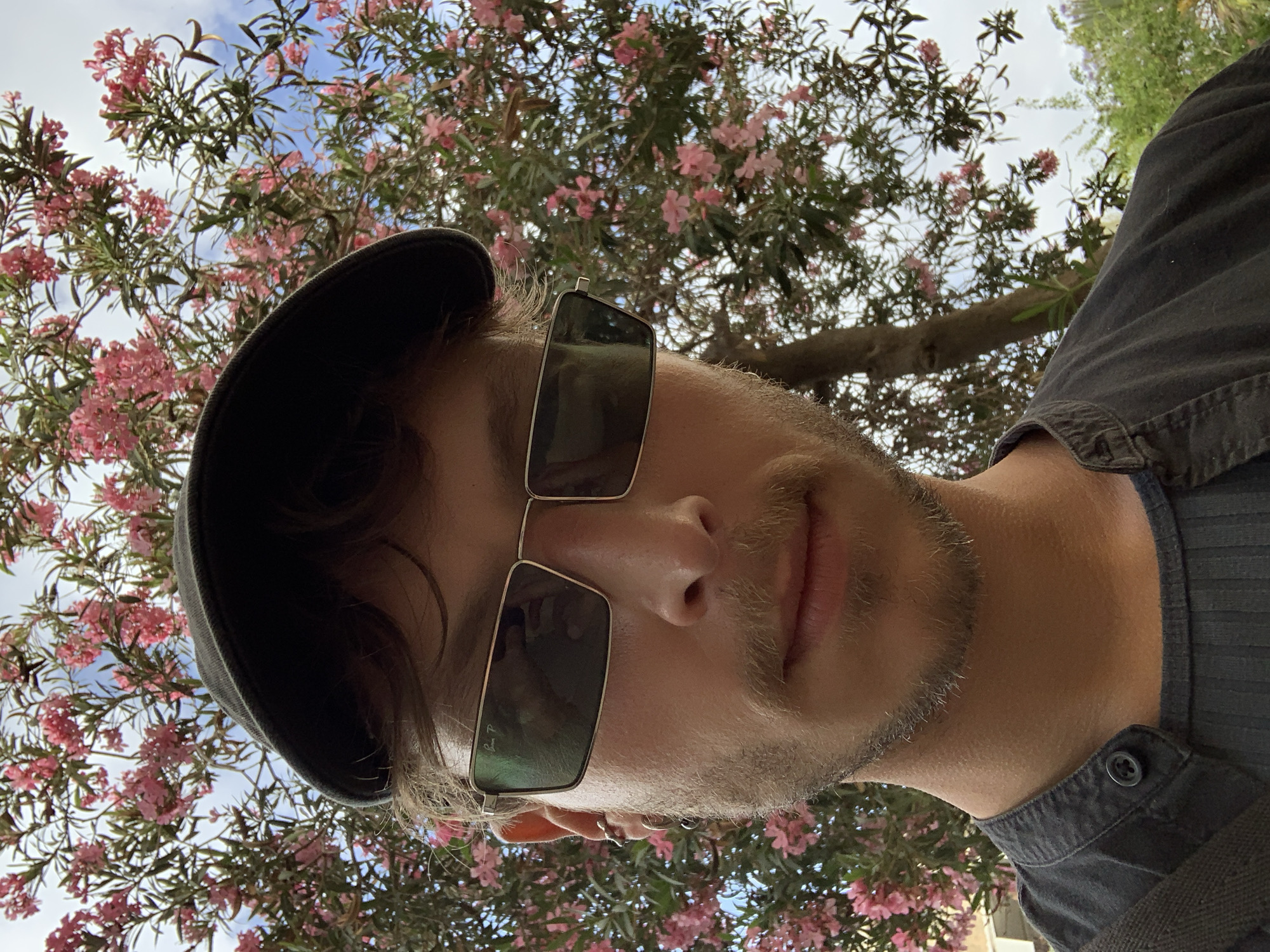 A selfie taken from a low angle shows a man with light skin and a beard wearing dark sunglasses and a dark cap. He is positioned in front of a tree with pink flowers, against a background of sky and foliage. The button of a dark-colored collared shirt is visible on the right side of the frame. The location appears to be Tel Aviv, Israel, based on the type of vegetation and architectural elements partially visible in the background.