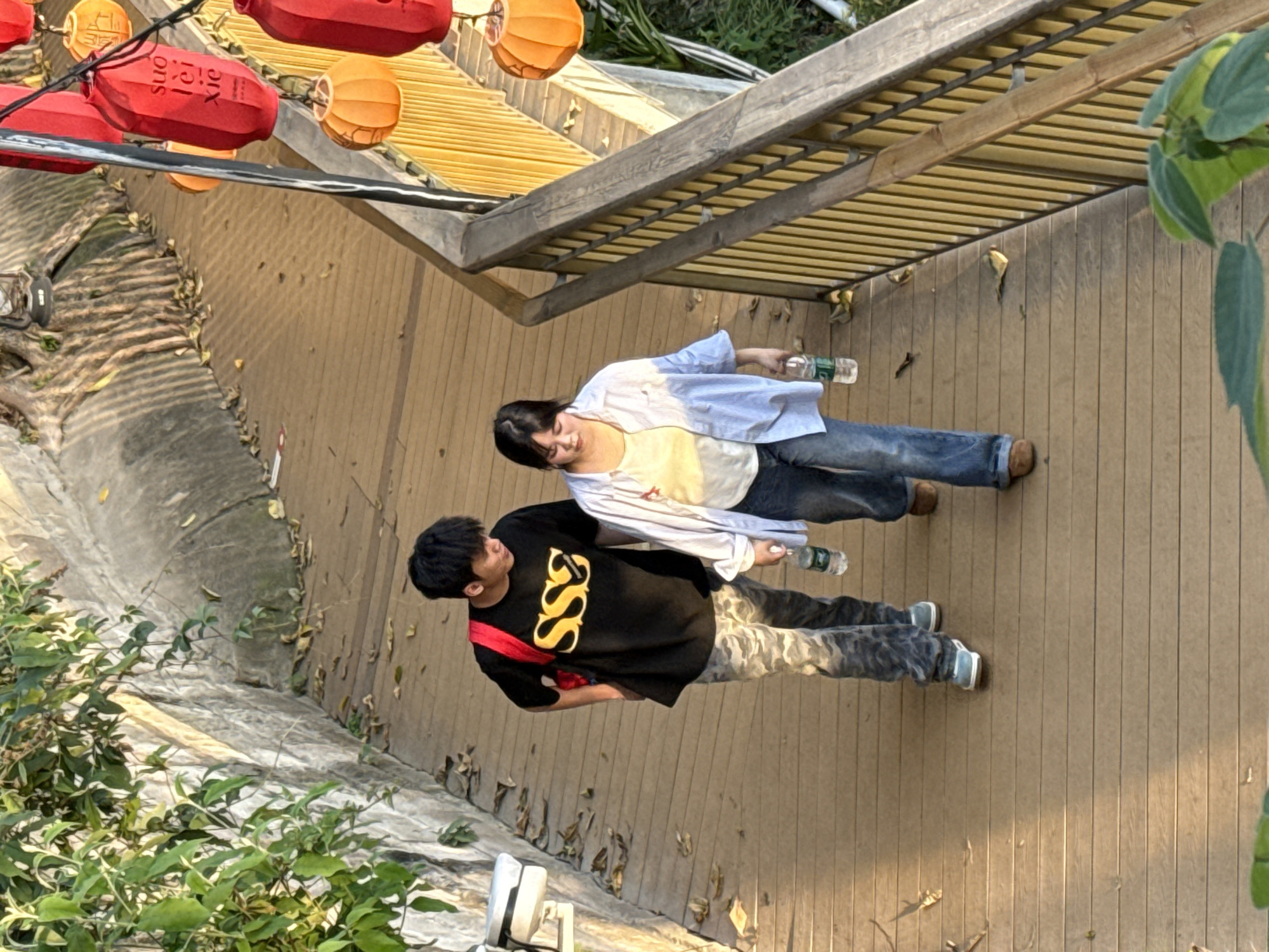 Observed outdoors in Yuzhong District, China, on a wooden boardwalk under red and orange lanterns, a woman in a yellow top, blue jeans, and a blue and white striped button-down shirt holds a water bottle. A man wearing a black t-shirt with gold script, camouflage pants, and a red strap walks nearby. The boardwalk is adjacent to a concrete and stone area with vegetation.