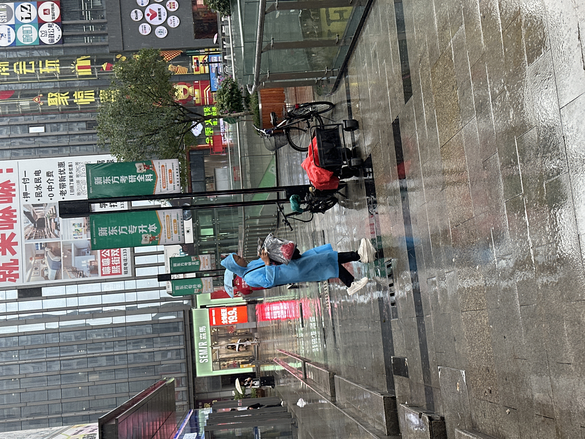 The image depicts an urban street scene in the Chongqing High-tech Zone, China, following a rain event, as indicated by the thoroughly wet and reflective dark grey tiled pavement. The sky appears overcast.
In the foreground, a person wearing a bright blue hooded raincoat, dark trousers, and white sneakers walks from left to right. They carry a transparent plastic bag containing red items in their left hand and a black shoulder bag on their right shoulder. Adjacent to them on the right, a black utility cart with red fabric or items rests near two bicycles—one electric and one traditional—parked next to a small tree planted in a square wooden planter.
In the midground to the left, under a covered walkway, a person with a white umbrella is visible, and another individual in dark attire stands further back. In the center-left background, a "SEM/R 森马" (Semir) store is visible with an illuminated red sign displaying "19.9".
The background features tall, modern buildings with extensive glass facades, reflecting the environment. Numerous commercial signs and billboards are prominently displayed on and around these buildings. Key visible signage includes "新东方专升本" (New Oriental Junior College to Undergraduate), "新东方考研全科" (New Oriental Postgraduate Entrance Exam All Subjects), "嘿嘛公寓" (Heima Apartment) with rental information, and a large red sign with Chinese characters and images of home interiors. Other illuminated signs bear Chinese characters such as "聚惊", "田园町", "LZ", "怡政公考", and "昂立教育". A person with a red umbrella is partially visible behind the main subject, near the right edge of the frame, further indicating the rainy conditions.