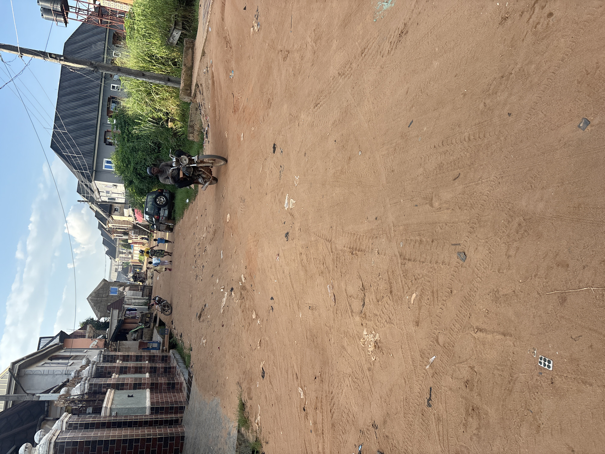 In Obosi, Nigeria, a dirt road traverses a residential area lined with multi-story buildings. The road surface is a loose, reddish-brown dirt, showing tire tracks and scattered debris, including plastic bags, wrappers, and what appears to be a discarded blister pack of pills. On the right side of the road, tall, green grass and vegetation grow, contrasting with the dry earth. A concrete drainage ditch runs along the right edge. In the background, a tall metal structure with two large water tanks is visible.
The scene is populated by several individuals. A man rides a motorcycle towards the viewer, his face partially obscured by a helmet and the bright sunlight. He is wearing dark clothing. Further up the road, a group of children and at least one adult are walking away from the camera. A parked motorcycle is visible near some buildings on the left side of the road. A dark-colored SUV is also parked on the left. The buildings on the left are constructed with brick and concrete, featuring decorative pillars. Buildings on the right are more modern, with dark pitched roofs and white-framed windows. The sky is partly cloudy.