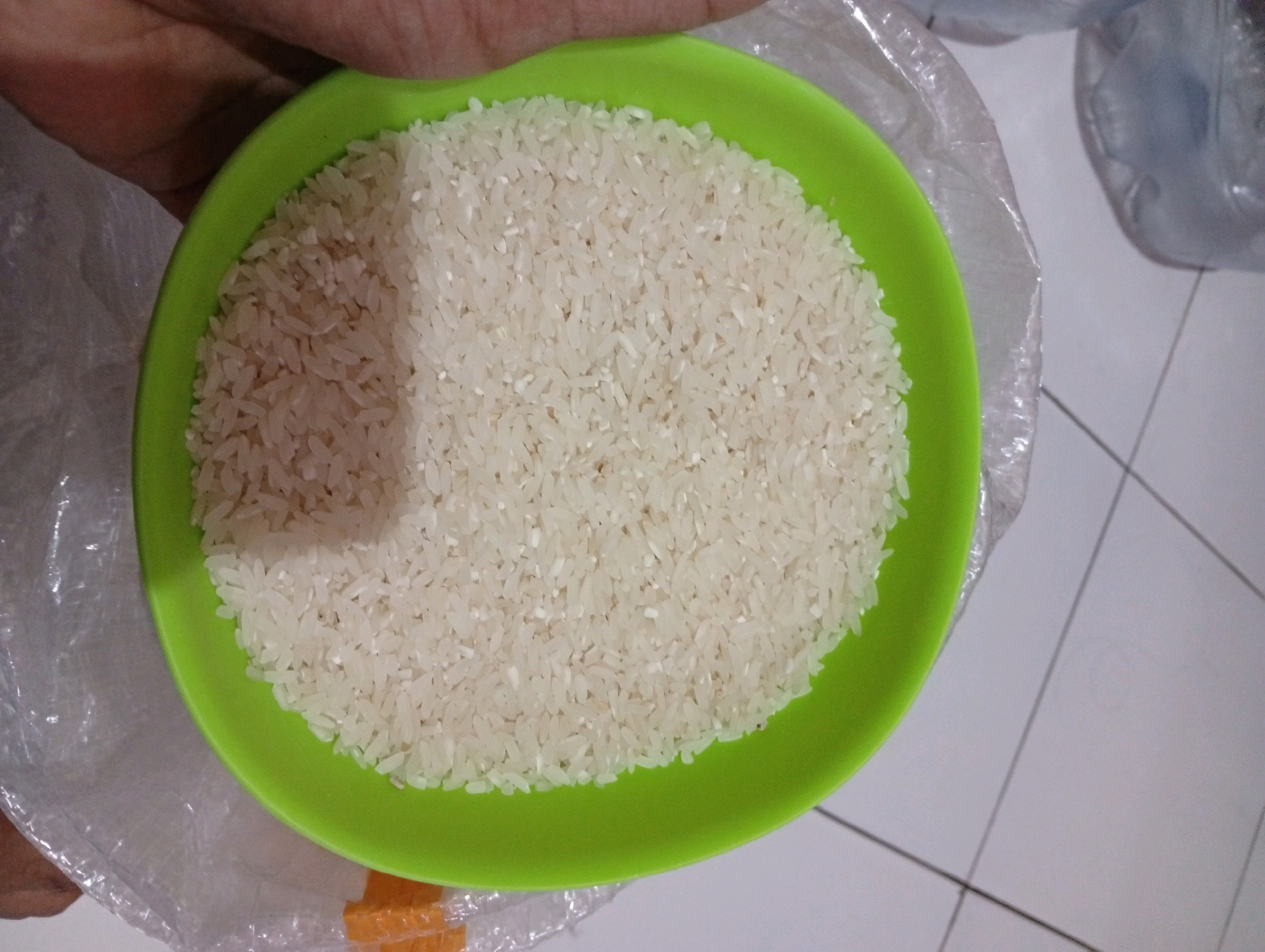 A bright green plastic bowl, filled with uncooked white rice, is held by a person's hand. The bowl is positioned slightly off-center to the left of the frame. The rice is a uniform color and texture, filling the bowl to the brim. The hand is partially visible at the top left corner of the image, with skin tone suggesting an adult. The background is out of focus and appears to be a tiled floor with some plastic wrapping. There is no identifiable information about the location in Tasikmalaya, Indonesia.