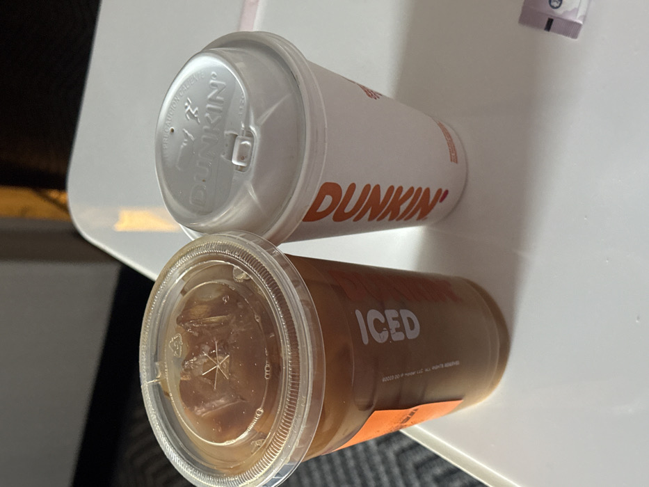 The scene, located in Unknown, Unknown, depicts two Dunkin' beverages resting on a smooth, white surface, likely a table. To the left, a clear plastic cup contains a light brown liquid with ice cubes visible through a clear, domed plastic lid. An orange adhesive label on the cup's side prominently features the word "ICED" printed vertically in white capital letters. To the right, a white paper cup displays the "DUNKIN'" logo in orange vertical text and is topped with a white plastic sip-through lid. A small, rectangular, partially visible packet, colored purple and white, is present on the white surface in the bottom right corner. The background is blurred, showing a dark area with a subtle patterned texture on the left and a warm, golden-yellow light or object on the right.