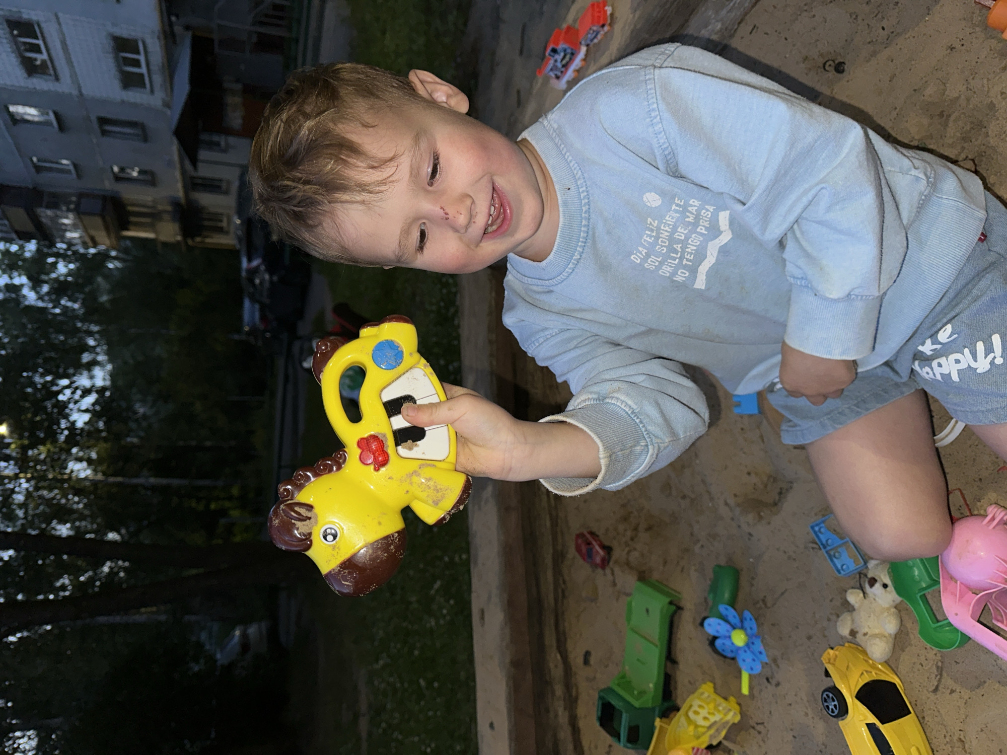 A young boy with blond hair and a light blue long-sleeved shirt is sitting in a sandbox in what appears to be a residential courtyard in Nizhny Novgorod, Russia. He is holding a yellow toy horse with a keyboard on its back. Several other toys, including a green truck, a yellow car, a small red car, a teddy bear, and a blue flower, are scattered in the sand around him. In the background, trees and a multi-story apartment building are visible. The lighting suggests it is late afternoon or early evening.