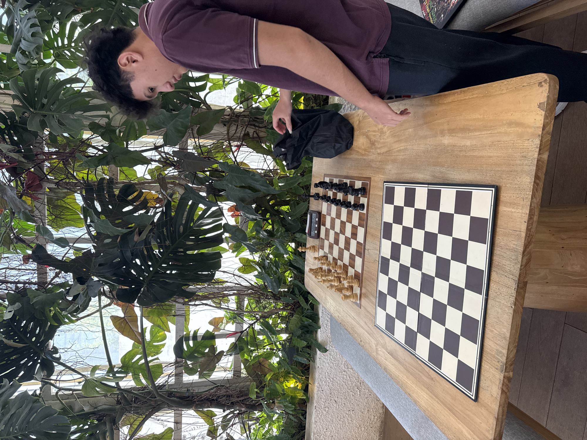 A young man stands at a wooden table, positioned in front of a lush tropical plant arrangement. He is wearing a maroon polo shirt and dark pants. On the table, there are two chessboards, one set up with chess pieces and a timer, and the other bare. The scene is bathed in natural light, suggesting an indoor setting with views of the outdoors. The location, North Kuta, Indonesia, is implied by the tropical plants.