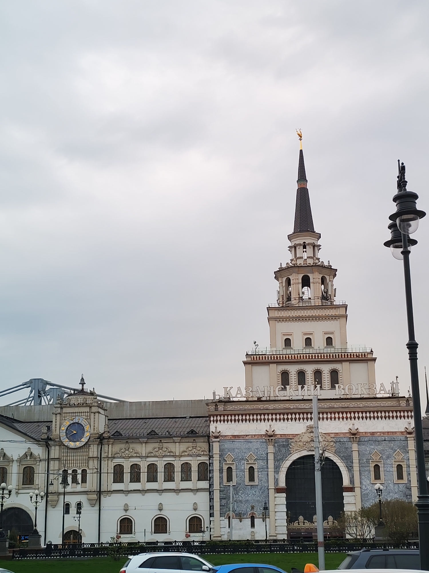 The image displays the grand architectural facade of Kazan Station in Moscow, Russia, under an overcast sky. The prominent building features a tall, slender spire on the right, capped with a distinctive golden, dragon-like finial. The spire rises from a multi-tiered tower with arched openings and decorative elements.
To the left of the main tower, a square clock tower showcases a large, ornate blue clock with golden Roman numerals. The station's lower levels exhibit a mix of white and patterned blue-grey stone facades, adorned with intricate carvings and arched windows. Above the central entrance arch, the Cyrillic text "КАЗАНСКИЙ ВОКЗАЛ" (Kazan Station) is clearly visible.
In the foreground, parts of several cars are visible, alongside a decorative black fence and green grass. A prominent black lamppost stands on the right edge of the frame. In the upper left background, part of a modern, metallic bridge structure can be seen. The weather appears cloudy, with a soft, diffused light suggesting daytime. No people or specific activities are distinctly visible in the static scene. The architecture conveys a sense of historical grandeur typical of major Russian landmarks.